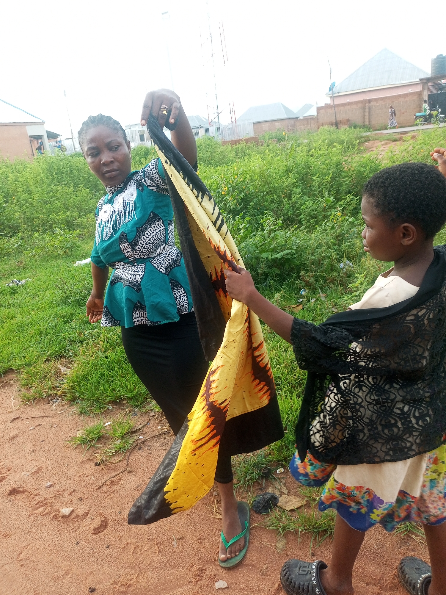 In Tempure, Nigeria, two individuals, an adult woman and a young girl, are positioned outdoors in an unpaved area with grassy vegetation bordering the scene. The woman, on the left, wears a teal and white patterned blouse and a black skirt, holding a yellow and black patterned fabric. The young girl, on the right, is dressed in a cream-colored top, a black lace-like overlay, and a colorful patterned skirt. She is also holding the fabric, suggesting a shared activity or interaction with the textile. The background reveals typical Nigerian urban housing, including brick walls and houses with pitched roofs, under an overcast sky. The presence of a motorcycle and people in the distance indicates a bustling, lived-in environment. The overall scene captures a moment of everyday life in a Nigerian community.