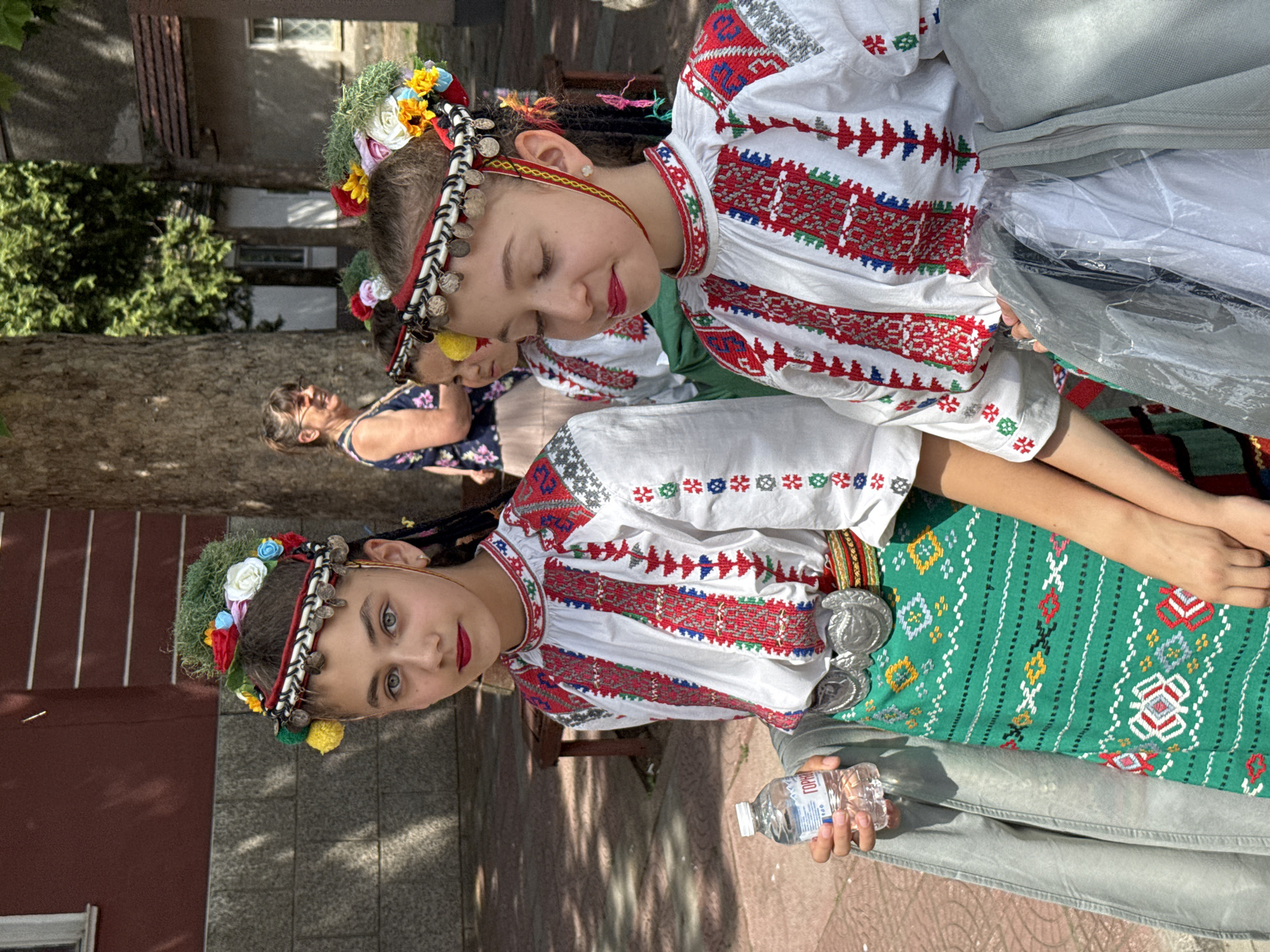 In Pordim, Bulgaria, two young girls in traditional Bulgarian folk costumes stand outdoors, positioned close to the viewer. The girl on the left faces forward, her eyes looking at the camera, while the girl on the right is angled away, her eyes closed in contemplation. Both girls wear intricately embroidered white blouses with red and blue patterns on the sleeves and collars, and brightly colored headpieces adorned with flowers and small silver coins. The girl on the left holds a bottle of water. In the background, another woman, seemingly an adult, stands partially obscured by trees, and a third child in a similar costume is visible behind the two foreground figures. The scene is set on a paved area with a brick pattern, under the shade of large trees, suggesting a public gathering or festival in Pordim.
