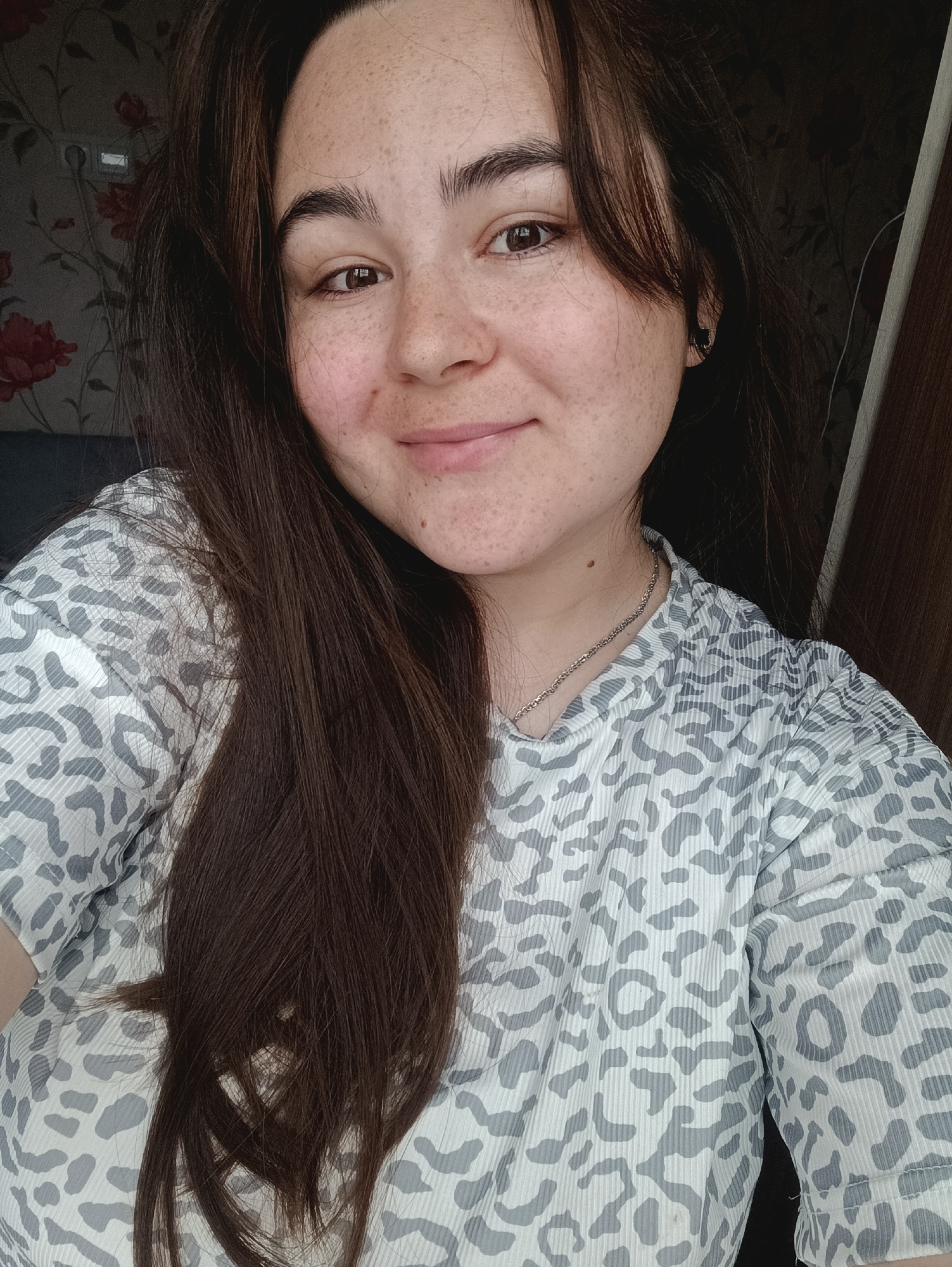 A young woman with long, dark brown hair and freckles is looking at the camera. She is wearing a gray and white leopard-print shirt and a silver chain necklace. She is indoors, in front of a floral-patterned wallpaper and a wooden door. The image was taken in Noviy, Russia.