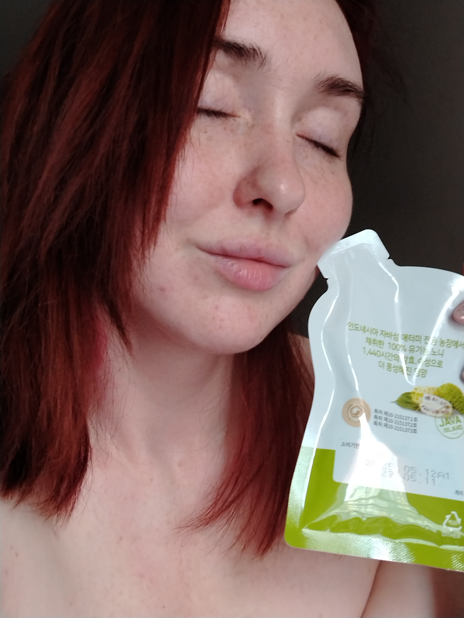A woman with reddish-brown hair and visible freckles is captured in a close-up shot, indoors in Izhevsk, Russia. Her eyes are gently closed, and her lips are slightly pursed, suggesting a moment of calm, contemplation, or appreciation. She holds a white and green flexible pouch close to her face, as if smelling or savoring the product inside.
The pouch features Korean text and an illustration of a noni fruit, depicted both whole and in cross-section. The visible text translates to "100% organic noni harvested from Atomy's entire farm on Java Island, Indonesia, with nutrients enriched by 1,440 hours of fermentation." The words "JAVA ISLAND" are prominently displayed on the packaging. Other Korean text includes "소비기한" (Expiration Date/Best Before), and three patent numbers: "특허 제10-2151371호", "특허 제10-2151372호", and "특허 제10-2151373호". Dates such as "2025.05.12" and "2027.05.11" are visible, followed by "까지" (until), likely indicating a best-before date. A recycling symbol labeled "OTHER" is also visible on the bottom right of the pouch.
The woman's hair frames her face and shoulders, and her skin shows a smattering of freckles across her nose and cheeks. The background is dark and indistinct, allowing the subject and the product to be the focal point. Soft lighting illuminates the scene, highlighting her features and the details of the packaging. Her overall expression conveys a peaceful and content interaction with the product.