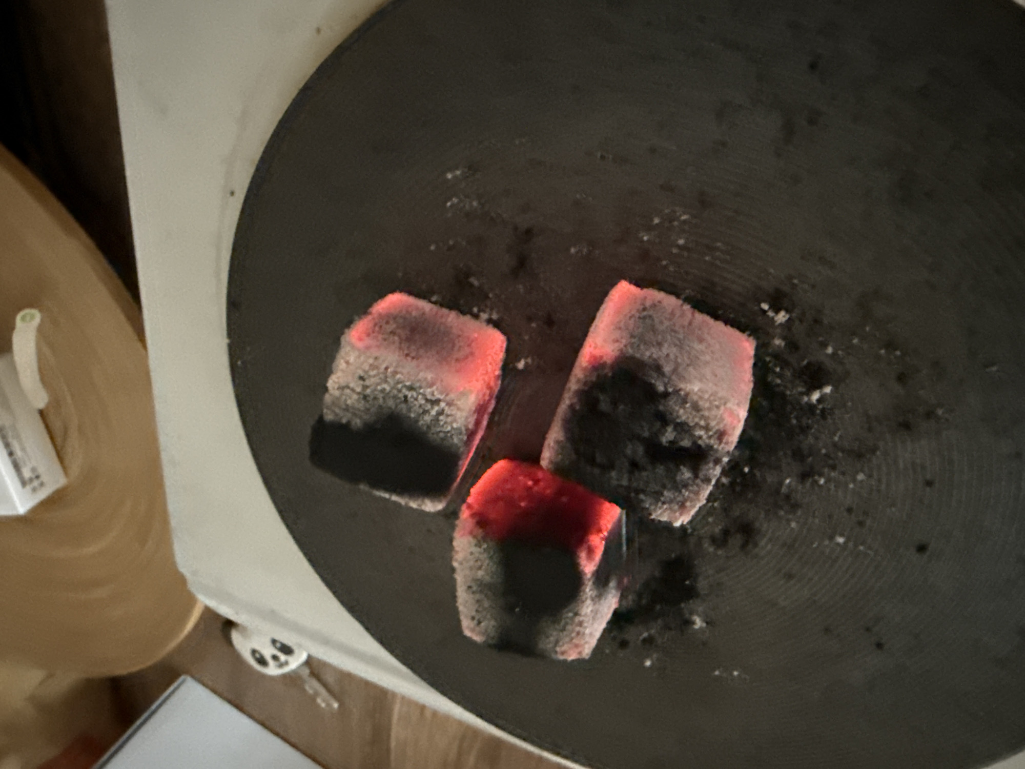 The image displays a close-up, top-down view of three actively burning coals resting on a dark, circular metal plate. The coals are roughly cubic in shape, glowing intensely with a vibrant orange-red color from their centers, while their outer layers are covered in a light grey ash. Small specks of ash and dust are scattered across the black metal surface, which features subtle concentric circular grooves.
The scene appears to be indoors, with various objects in the background, mostly out of focus. To the left, a curved, light brown surface, possibly wood, is visible. Adjacent to it, a white object with a small green tip can be seen. In the bottom left, a corner of a white box with faint, unreadable black text is present. To the bottom right, a silver key with a distinctive white panda-shaped keyring peeks out from beneath a white surface. The prominent glow of the coals suggests the photograph was taken in a dimly lit environment, possibly at night or in the evening. The coals are likely being prepared for use, such as with a hookah. The location is Varna, Bulgaria.
