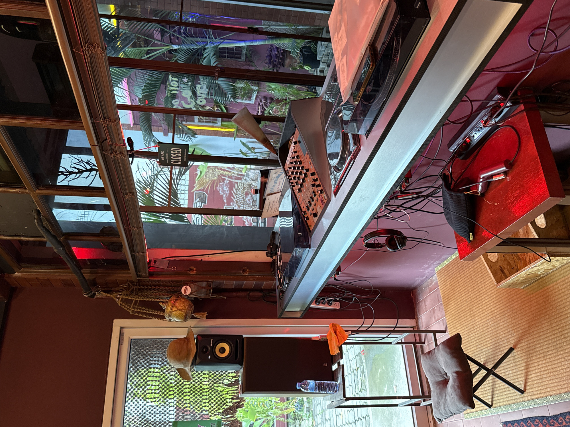 This image depicts the interior of a DJ setup in a room with a brown and red color scheme, likely located in Bangkok, Thailand based on the tropical vegetation visible through the window. A DJ console with turntables and a mixer is prominently featured, wired with numerous cables. A power strip is visible underneath the console. Red overhead lighting casts a glow on the equipment and surrounding surfaces.
To the side of the DJ setup, there's a window overlooking greenery. Below the window, a shelf holds a speaker and a bottle of water. A folding chair with a gray cushion sits on a floor covered with a woven mat. Above the window, a decorative fishing net hangs from the ceiling. A "CLOSED" sign is hanging near the window, partially obscured by the interior decor.