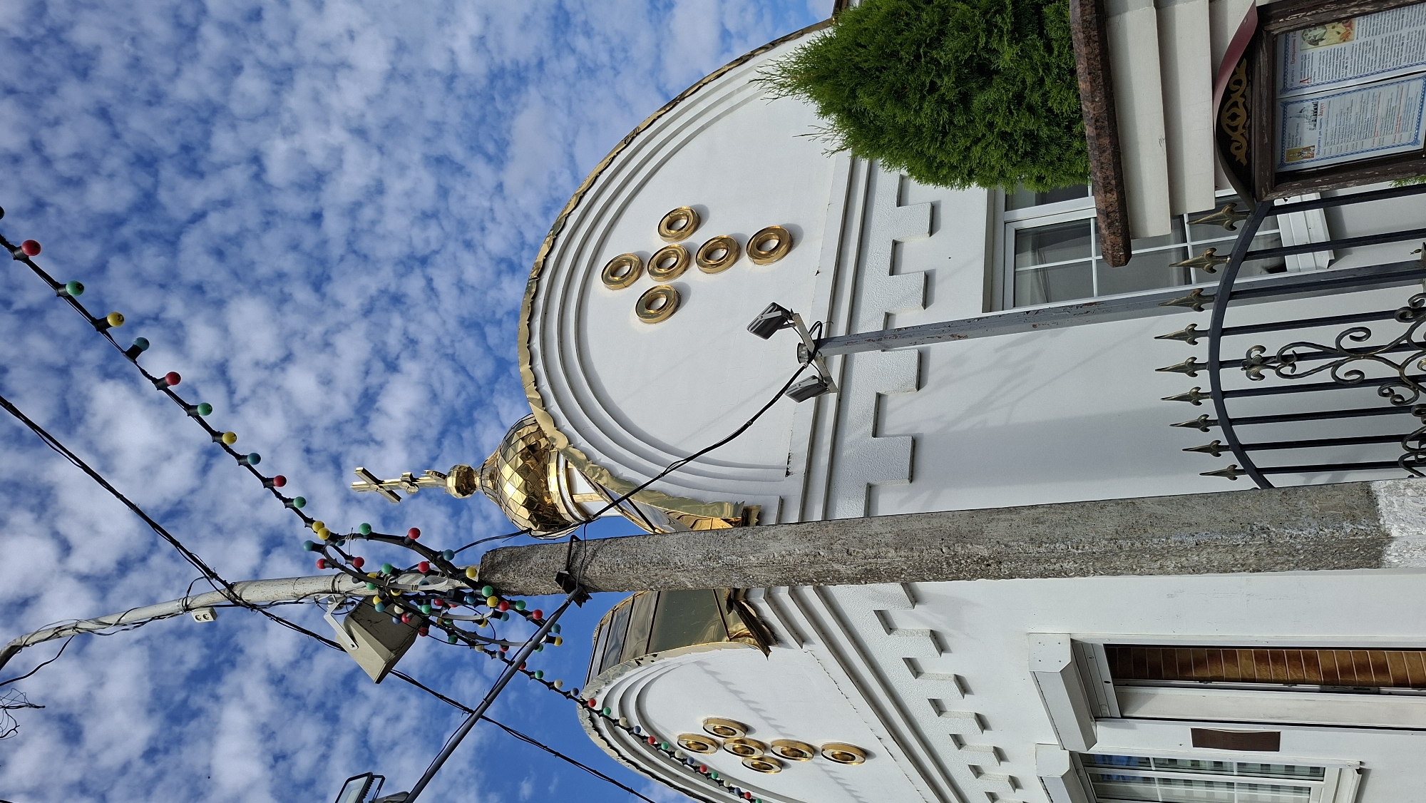 The image shows the exterior of a white building with gold-trimmed architectural features and a gold dome topped with a cross. The building appears to be a church, possibly Orthodox given the architectural style. It is decorated with colorful string lights that hang from wires overhead, which also carry electrical cables. A concrete utility pole stands in the foreground, also adorned with some of the string lights and other electrical equipment. The sky is blue with scattered white clouds. A black wrought-iron fence is visible in front of the building, and a lush green tree is to the right. A wooden sign with text and images is also present near the fence. The overall scene suggests a bright, partly cloudy day in a city environment, possibly Pastavy, Belarus, given the context.