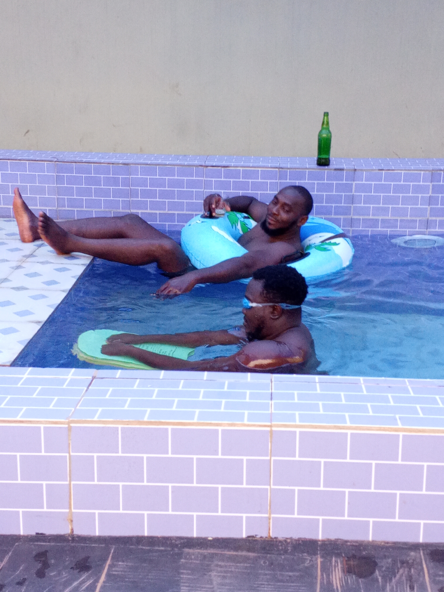 Two Black men are in a tiled swimming pool. One man is floating on a light blue inner tube with white designs, his legs extended out of the water and resting on the tiled edge of the pool. The second man is in the water, wearing swimming goggles, with his hands on a light green kickboard. A green glass bottle stands on the tiled pool edge behind the man on the inner tube. The location is suggested to be Ankpa, Nigeria, but there are no definitive location identifiers in the image.