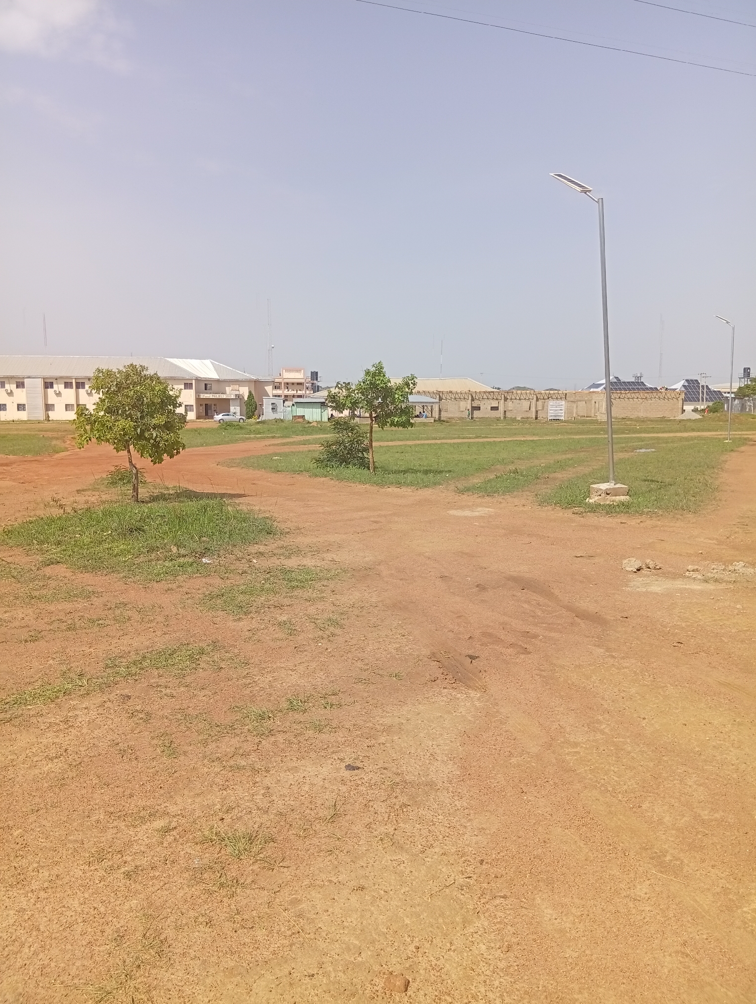 The image shows an outdoor scene with a dusty, reddish-brown dirt path in the foreground. Scattered patches of green grass and a few small trees are visible. In the background, there are several buildings, including a long, light-colored structure with a white roof, an industrial building with solar panels on the roof, and a taller, multi-story building in the distance. A modern street lamp with a solar panel is positioned on the right side of the frame. The sky is a pale blue with some hazy clouds. There are no people or animals visible in the image. The setting appears to be an undeveloped or construction area within an urban or semi-urban environment in Gwafan, Nigeria.
