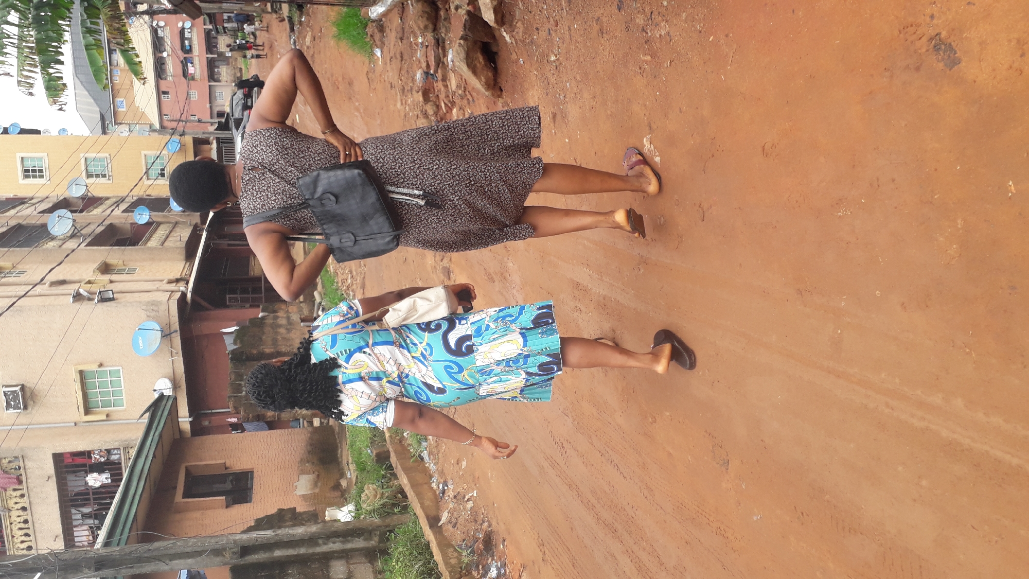 Two women are walking on a dirt road in Nkpor, Nigeria. The woman in front wears a blue and white patterned dress and black sandals, carrying a white bag. The woman behind her wears a brown patterned dress, brown sandals, and a black shoulder bag. They are walking away from the camera, and the road they are on is reddish-brown. Buildings with satellite dishes are visible in the background. Green vegetation borders the road.