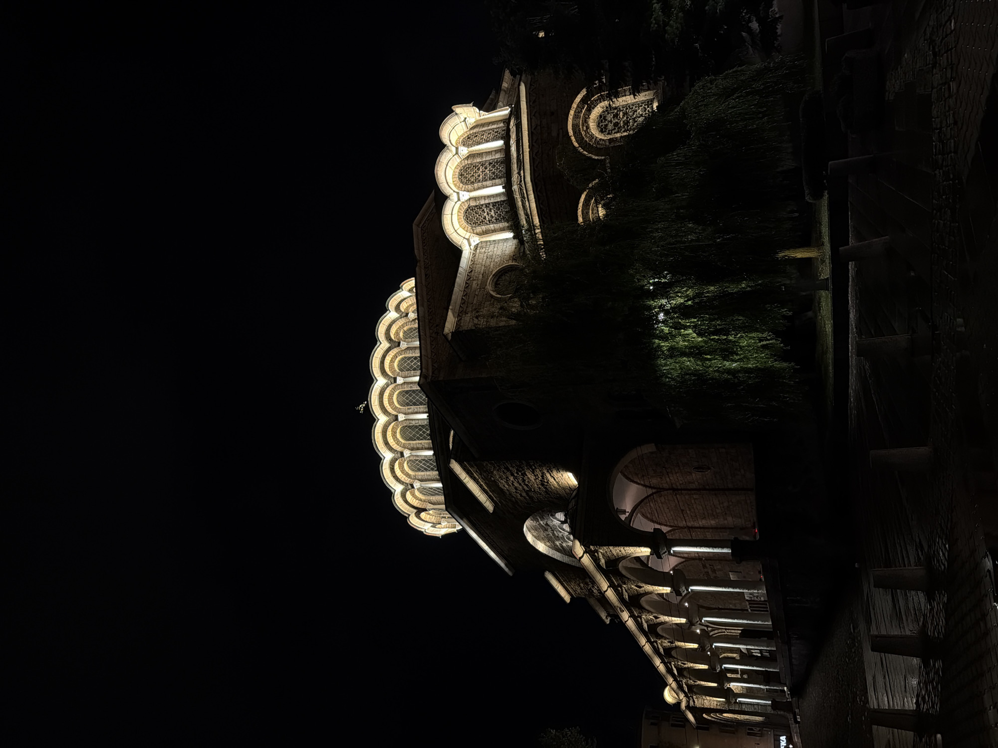 This media file captures a dramatic night view of an ornate building in Sofia, Bulgaria. The image is oriented sideways, but when viewed correctly, it depicts the building rising into a dark, starless sky.
The main subject is the architecturally distinctive building, illuminated by warm, artificial lights. Its facade features multiple levels of intricate design. The upper sections are particularly notable for their series of scalloped arches, each housing decorative windows with patterned grilles that glow brightly. These patterns suggest a style with Moorish or Byzantine influences.
Below these ornate upper windows, the building continues with more archways and sturdy, stone or brickwork construction. Elements like columns or pilasters are also lit, casting highlights and shadows that emphasize the building's texture and depth. Dark, dense foliage from trees or shrubs is visible on the right side of the image (when oriented correctly), partially obscuring parts of the structure and adding to the nocturnal atmosphere. The foreground, particularly on the lower right, suggests a paved area or street running alongside the building, with some dark, horizontal structures visible.
There are no people visible, and no specific event is taking place, making it a static architectural study. The time of day is clearly night, with the artificial lighting creating a striking contrast against the black sky.
A notable detail, revealed in a closer crop, is a partial sign on the building's lower level that reads "ОПТИКА" (Optica), indicating an optics shop or related business. This suggests the building is either commercial or a mixed-use historic structure. The overall impression is one of grandeur and historical significance, beautifully highlighted under the night sky.