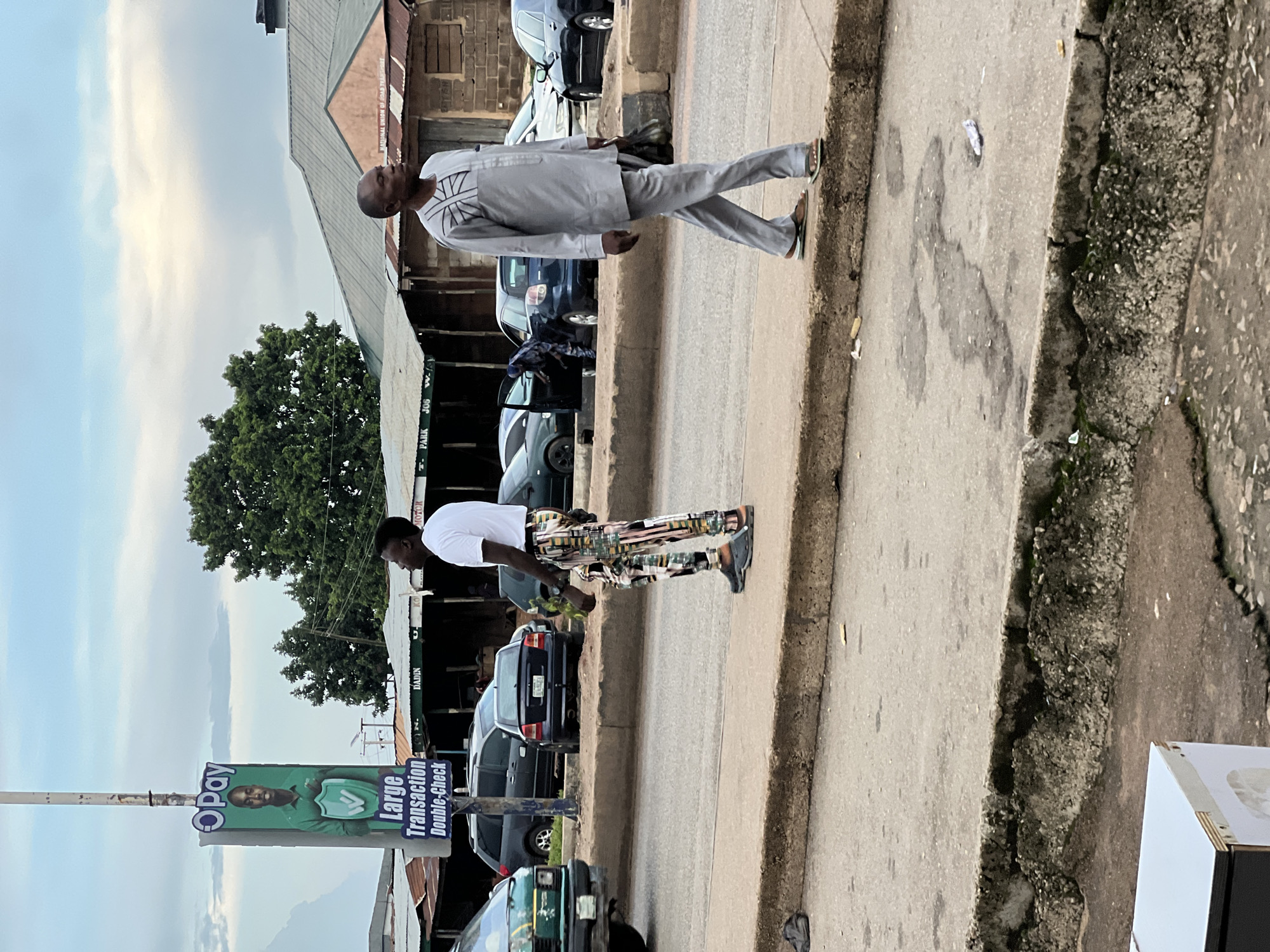 Two men are standing on a sidewalk next to a street. The man on the left is wearing a white t-shirt and patterned pants, and the man on the right is wearing a grey traditional outfit. In the background, there are cars and buildings. A large advertisement for "OPay" is visible on a pole. The environment appears to be an urban setting with moderate pedestrian and vehicle traffic. The sky is overcast.