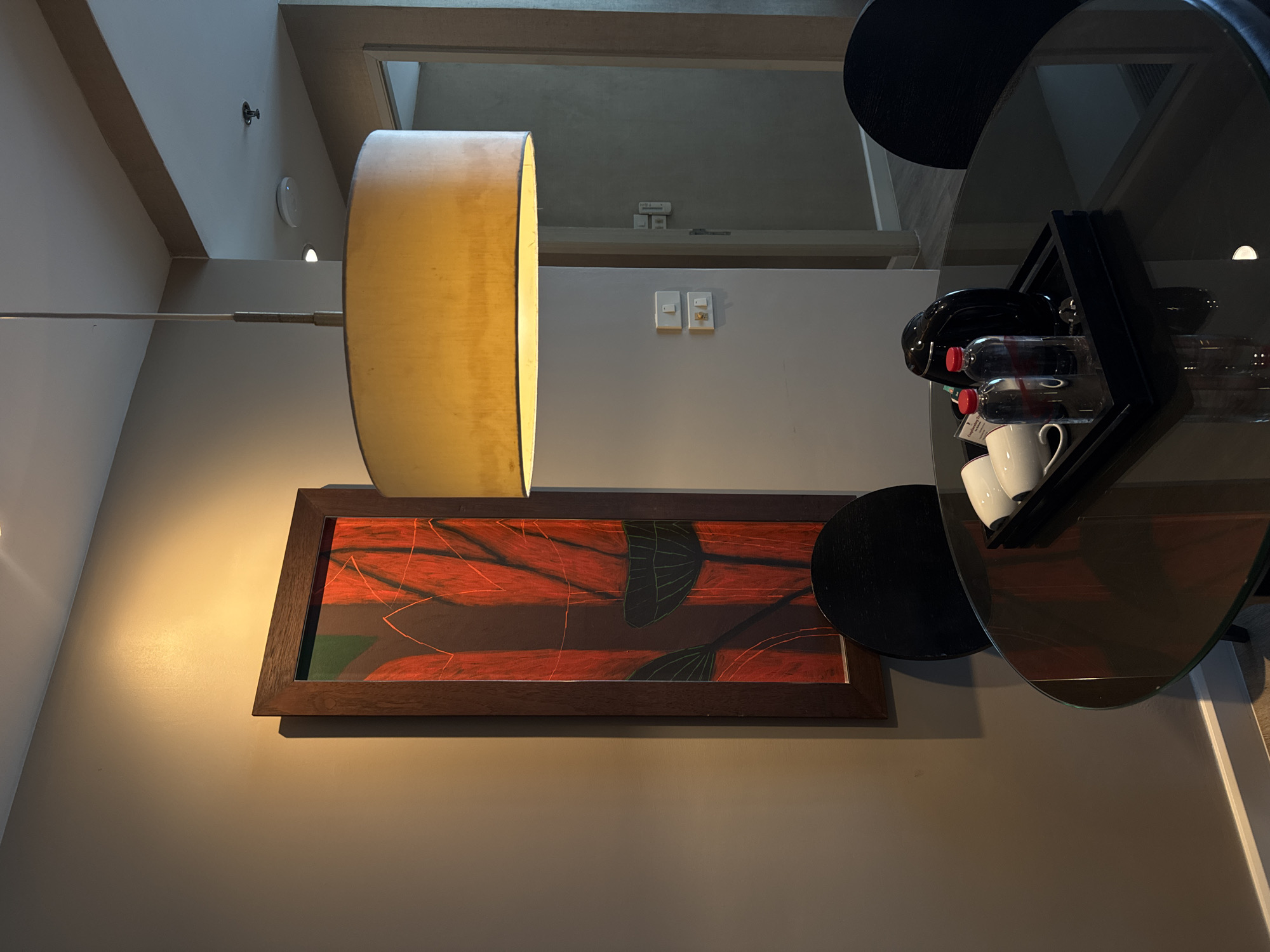 The image depicts an interior scene in Bangkok, Thailand, likely a hotel room or a serviced apartment, featuring a small dining area. In the foreground, a round, dark glass table with a metallic base is visible. On the table, a black tray holds two clear plastic water bottles with red caps, two white ceramic mugs, and a black electric kettle. Two black chairs with rounded backs are positioned around the table.
To the left, a large, rectangular framed artwork hangs on a tan-colored wall. The artwork features an abstract design with dominant red and dark green colors, with a dark wooden frame. A large, drum-shaped pendant lamp with a cream-colored fabric shade hangs from the ceiling, casting a warm yellow light.
In the background, a doorway leads into another room, with a light gray wall on the right side of the frame. Electrical outlets and light switches are visible on the wall to the left of the doorway. The floor appears to be light-colored laminate or wood. The overall ambiance is modern and functional.