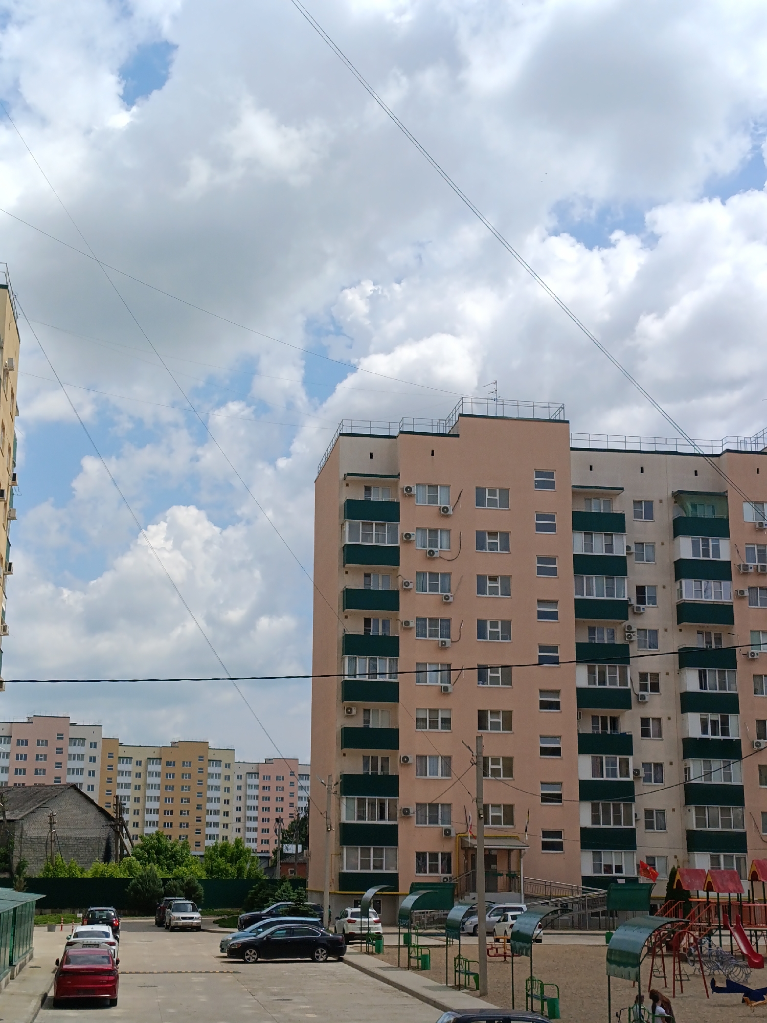 The image shows a residential area in Yablonovskiy, Russia, under a partly cloudy sky. A tall, peach-colored apartment building with dark green accents dominates the right side of the frame. The building is equipped with numerous air conditioning units and windows. In the foreground, a playground is visible, with partial views of play structures. A street with parked cars runs horizontally across the lower half of the image. In the distance, several other apartment buildings of varying colors can be seen behind some greenery and a smaller, older building. Power lines crisscross the sky above the buildings.