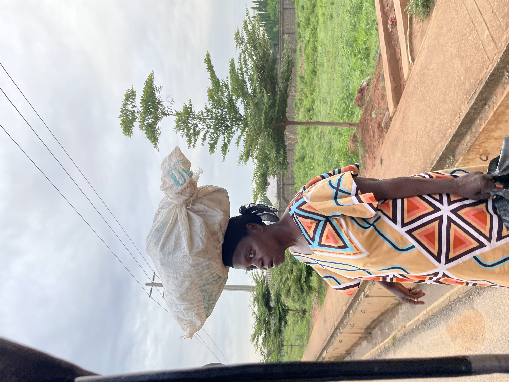 A woman, seen in a medium shot from the chest up, walks with a large white sack balanced on her head. She wears a black headscarf and a colorful dress with a geometric pattern of triangles in shades of orange, teal, and black on a yellow background. Her expression is focused and her mouth is slightly open. She is on a paved path next to a road with a concrete barrier. Behind her, there is a field of green grass, a small tree, and a concrete wall. Power lines and a utility pole are visible in the cloudy sky. The scene appears to be outdoors in an urban or semi-urban environment.