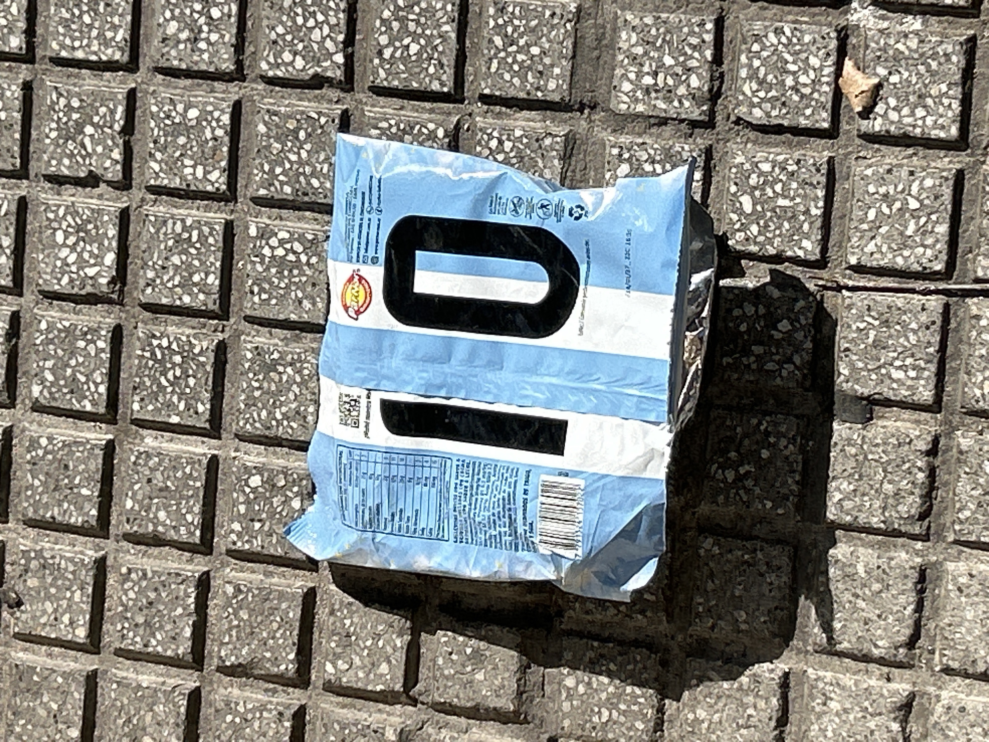 A crumpled, light blue and white striped plastic bag, resembling a snack wrapper, lies on a grey, textured, square-tiled pavement. The bag is emblazoned with large, black Arabic numerals "10" in the center. White text and a barcode are visible on the left side of the bag, along with a red and yellow circular logo. The pavement tiles are weathered and appear to be part of a public sidewalk or plaza in Buenos Aires, Argentina. Sunlight casts a distinct shadow from the bag onto the tiles. A small, dry brown leaf rests on one of the pavement tiles to the lower right of the bag.