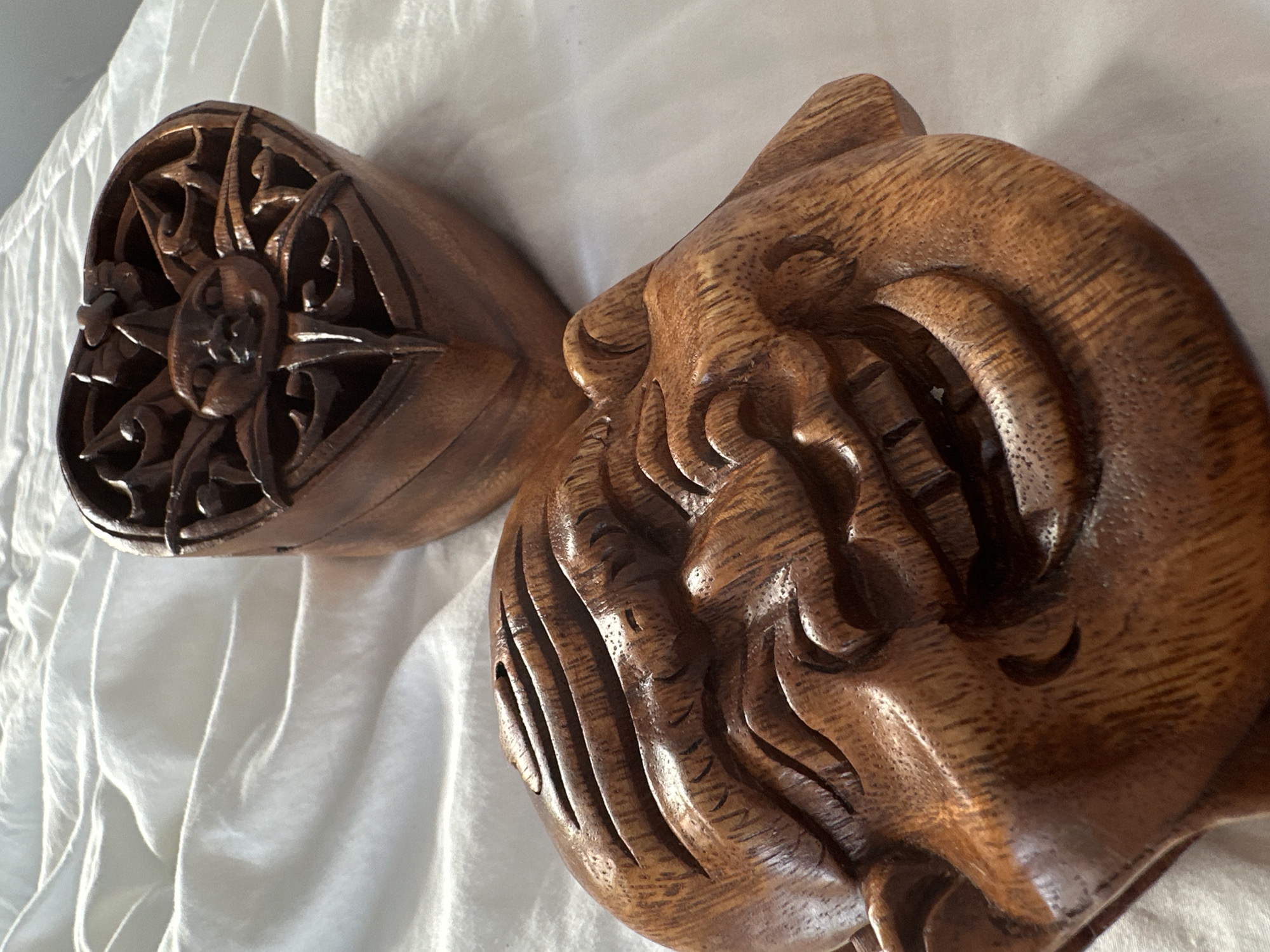 The image displays two intricately carved wooden objects resting on a soft, white fabric surface with gentle folds, likely indoors under natural light. The objects are made of dark brown wood with visible grain and a polished finish, which accentuates the carvings and creates soft highlights and shadows.
The object on the left features a deeply carved, circular motif at its end, resembling a stylized sun or star with radiating, ornate patterns and swirling tendrils. This piece appears to be a decorative element, possibly a finial or part of a larger wooden structure.
The dominant object on the right is a detailed relief carving of a stylized face, possibly depicting a mythical figure, deity, or a highly decorated human. It features prominent, defined eyes, a nose, and a mouth area, all integrated within a flow of elaborate, wavy, and deeply incised lines that form a headdress, hair, or abstract ornamental elements around the face. The craftsmanship highlights the texture and depth created by the carving.
There is no specific activity or event occurring, other than the display of these artistic pieces. The objects' intricate style and material are characteristic of traditional craftsmanship found in regions like North Kuta, Indonesia, known for its rich heritage of wood carving, particularly Balinese art. No text is visible in the media file.