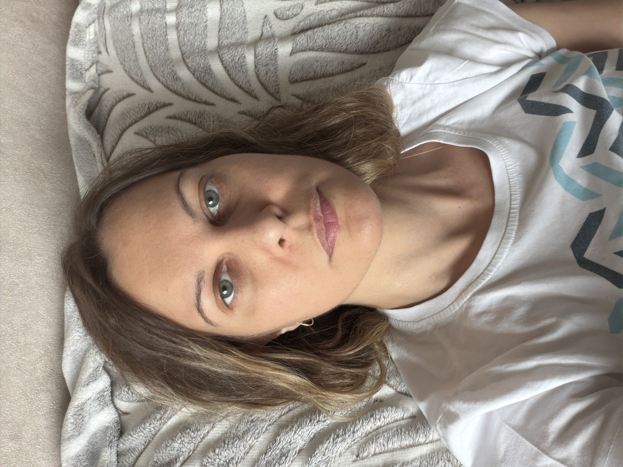 A woman is lying on a textured grey blanket. She has light brown hair and light colored eyes, and is wearing a white t-shirt with a geometric pattern on the sleeve. The scene does not indicate a location in Izhevsk, Russia or any specific city.