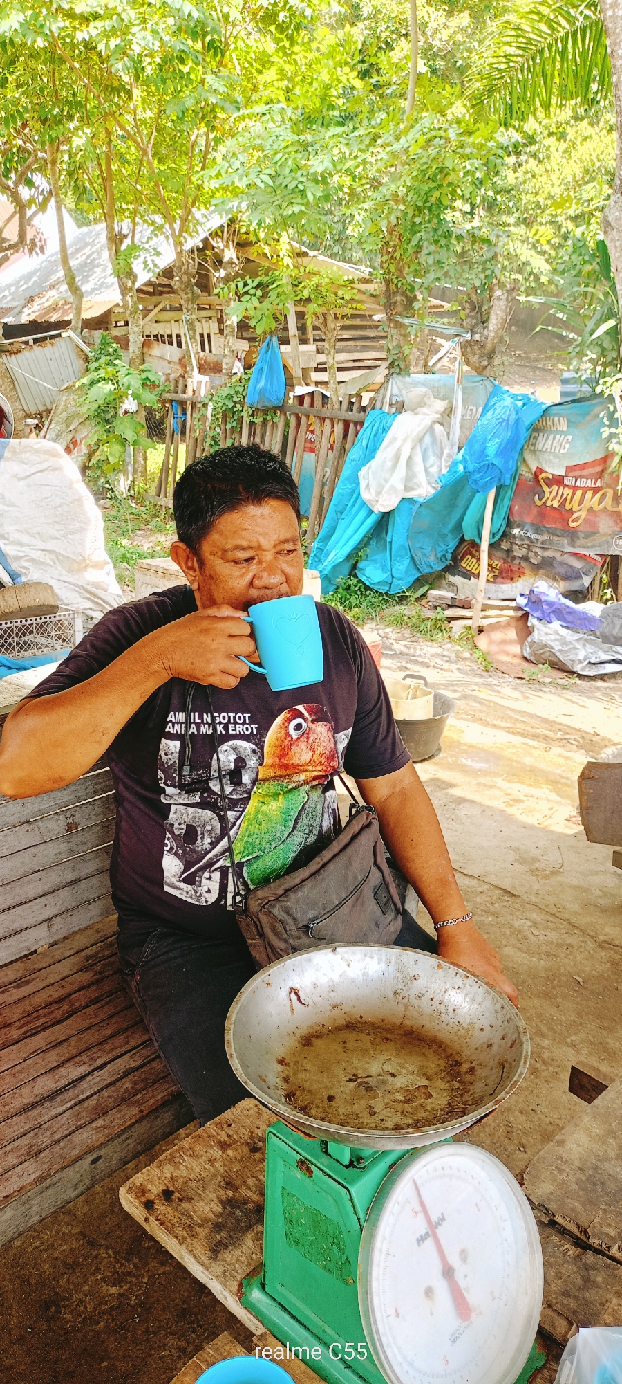 This media file captures a candid, eye-level shot of a man drinking from a light blue mug outdoors in Lhokseumawe, Indonesia. The man, who appears middle-aged with dark hair, is seated on a wooden bench. He wears a dark t-shirt featuring a prominent, colorful graphic of a parrot and text that reads "AMI IL N GOTOT ANI A MAK EROT." A dark cross-body bag rests on his lap. He holds the mug with both hands, his eyes cast downwards as he sips.
To his right, an old, green mechanical weighing scale with a tarnished silver pan is visible, resting on a wooden surface. The scale's dial shows markings, and the brand "Hà Nội" is faintly discernible. The background is lush with vibrant green foliage and trees, indicating a tropical or rural setting. Simple wooden and corrugated metal structures are partially visible through the greenery. Several blue tarpaulins or bags hang or are draped in the background, alongside a white bag and a weathered banner with text including "MENANG" and "KITA ADALAH SURYA." The bright, natural daylight suggests it is daytime. The overall scene conveys a sense of everyday life in a rustic outdoor environment. A "realme C55" watermark is present in the lower right corner.