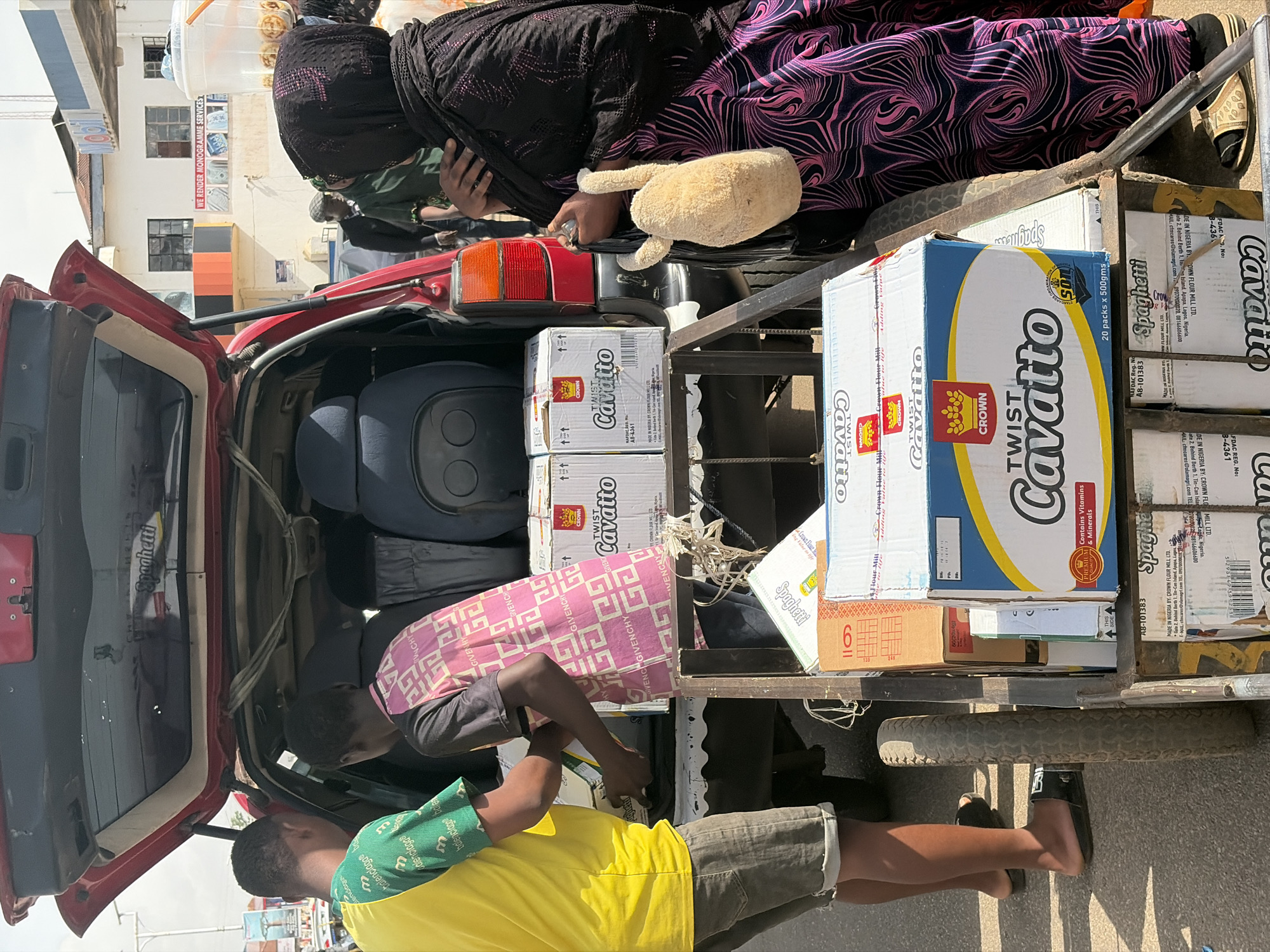 The image shows a busy scene in Jos, Nigeria, with several people and objects in and around a red vehicle. Two young boys are visible in the foreground, one wearing a yellow t-shirt and grey shorts, the other in a pink and white patterned long-sleeved shirt and grey shorts. They appear to be assisting or interacting with the contents of the vehicle.
The back of the red vehicle is open, revealing a load of boxed goods, primarily "Cavatto" brand spaghetti. The boxes are stacked high, suggesting a commercial transaction or delivery. One person, partially obscured, is wearing a black head covering and a purple and black patterned dress. They are holding a beige plush toy.
In the background, a building with windows and signage is visible, including text that reads "WE RENDER MONOGRAMME SERVICES." The lighting suggests it is daytime, likely a sunny day given the strong shadows. The overall impression is one of active commerce or logistics taking place on a street in Nigeria. The visible text on the boxes includes brand names, product information like "Spaghetti," "20 packs x 500gms," and regulatory details such as "NAFDAC Reg. No." and "MADE IN NIGERIA BY CROWN FLOUR MILL LTD." There is also visible text on the clothing of the boy in the foreground, reading "balenciaga" and "GIVENCHY".