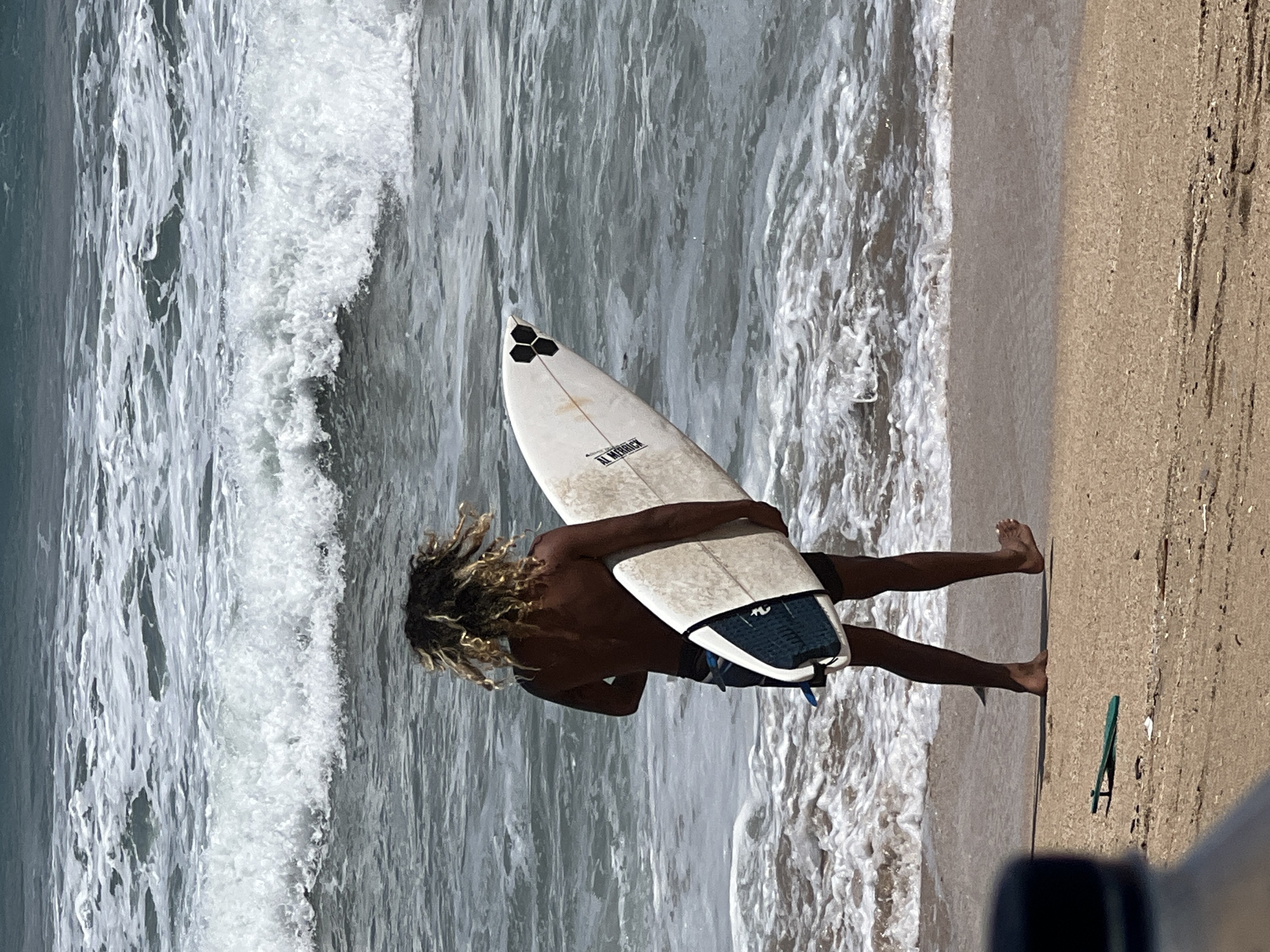 A surfer with curly, blonde-streaked hair walks on a sandy beach toward the ocean, carrying a white surfboard. The surfboard has "AL Merrick" written on it and features a black three-hexagon design at the tail. The surfer is wearing dark swim trunks. The ocean waves are breaking gently, and the water is a deep blue-gray. The beach is a light brown sand. In the foreground, a blurry dark bottle cap is visible on the left. A small green object, possibly a fins accessory, lies on the sand to the left of the surfer's feet. The overall scene suggests a surfing activity in Kuta, Indonesia.