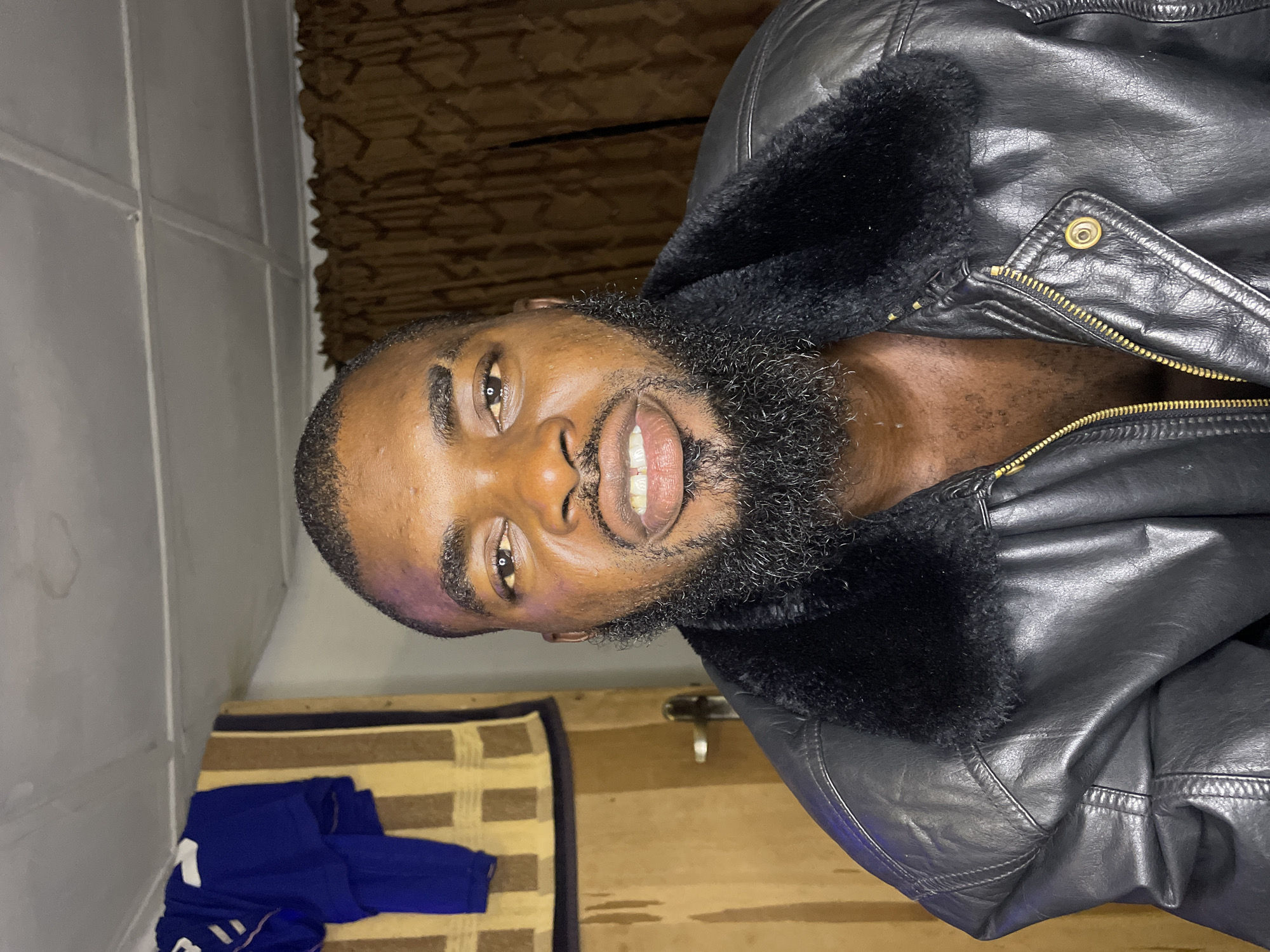A male individual with dark skin, a full black beard, and short dark hair is centrally positioned, facing the camera directly. He is wearing an unzipped black leather jacket featuring a black faux fur collar, revealing a portion of his chest. His mouth is slightly open, showing his upper and lower teeth.
The background on the left side reveals a light brown wooden door with a silver-colored doorknob, above which a blue garment with white markings and a vertically striped brown and beige fabric are hanging. On the right side, a brown curtain with a subtle pattern is visible. The ceiling, painted light gray, exhibits multiple circular water stains and visible panel lines. The scene is an indoor setting, reportedly located in the city of Jos, Nigeria.