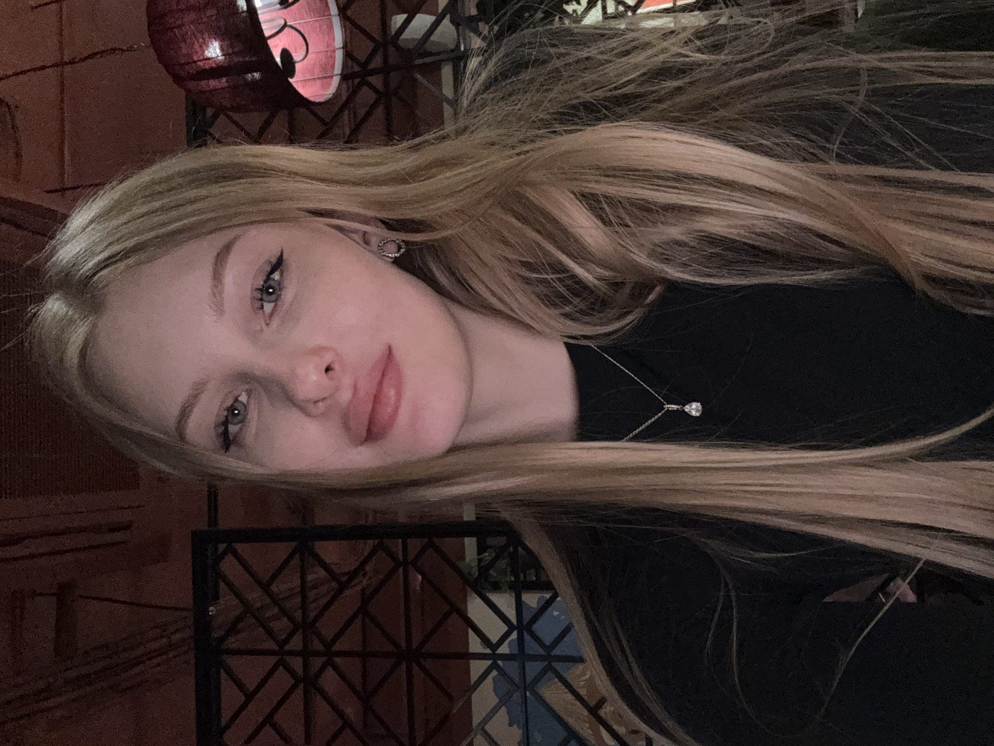 A close-up photograph depicts a young adult female from the chest up, positioned centrally and looking directly at the camera. She has long, straight blonde or light brown hair that falls over her shoulders. Her eyes are light in color, likely blue or grey, and are accentuated with black winged eyeliner. She wears a black long-sleeved top, a silver necklace with a single teardrop-shaped clear stone pendant, and small circular textured earrings. Her lips are slightly parted. The ambient lighting is dim and warm, casting a reddish hue on the scene.
The background is out of focus, revealing elements consistent with an indoor establishment, possibly a restaurant or bar. To the left, a black metal decorative screen with a geometric lattice pattern is partially visible. To the right, a red fabric lampshade, possibly pleated or textured, hangs from the ceiling, emitting a warm glow. Above these elements, a reddish-brown textured ceiling or wall surface is discernible, with subtle indications of pipes or structural elements.
The specific location within the city of Perm, Russia, is not ascertainable from the visual evidence provided.