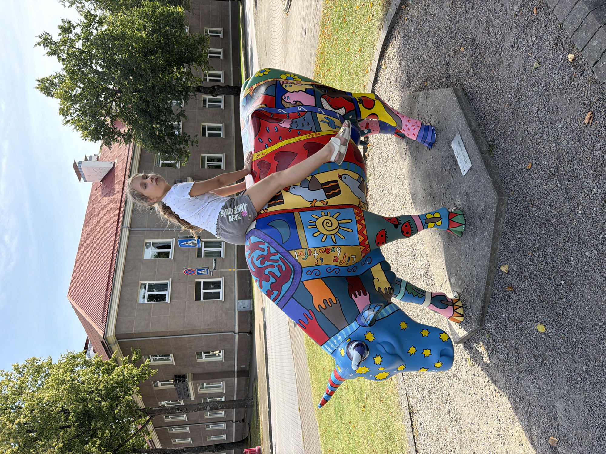 A young girl with blonde hair pulled into a ponytail sits on a colorful, life-sized sculpture of a cow, which is painted with various patterns and images, including stars, ladybugs, and abstract figures. The sculpture has a unicorn-like horn. The girl is wearing a white t-shirt and grey shorts. She is looking towards the camera with a neutral expression. The cow sculpture is positioned on a grey, hexagonal concrete base in a paved area covered with gravel. Behind the sculpture is a building with a red tiled roof and several windows, and trees with green foliage. The scene appears to be outdoors on a sunny day. This cow sculpture, titled "Dzīve ir skaista" (Life is Beautiful), is part of the cow parade art installations in Ventspils, Latvia, a city known for its numerous public art pieces, particularly along the Venta River embankment and throughout the city center.