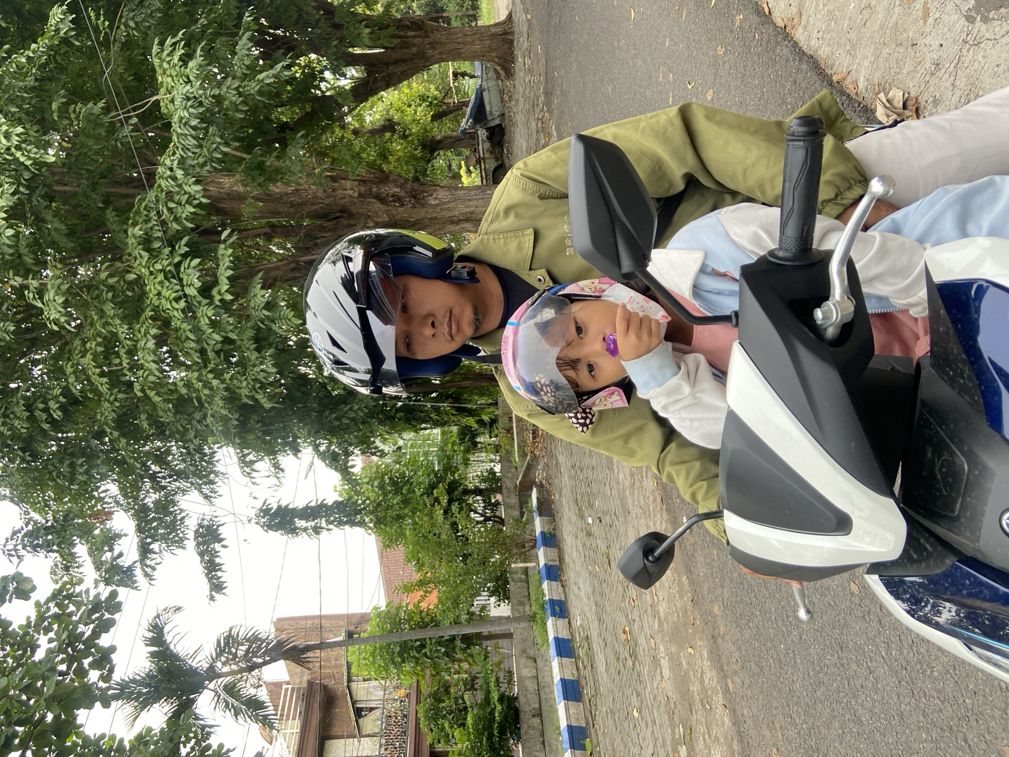 A medium shot captures a man and a young child on a parked scooter. The man, wearing a green jacket and a black motorcycle helmet with a blue visor, is positioned behind the child. The child, wearing a pink helmet and a light blue jacket, is seated in front of the man, facing the camera, with their finger in their mouth.
The scooter is partially visible, showing the handlebars, mirrors, and part of the front panel. The scooter is parked on a paved road with a blue and white striped curb.
The background includes trees, houses, and other buildings. The location is Surabaya, Indonesia.