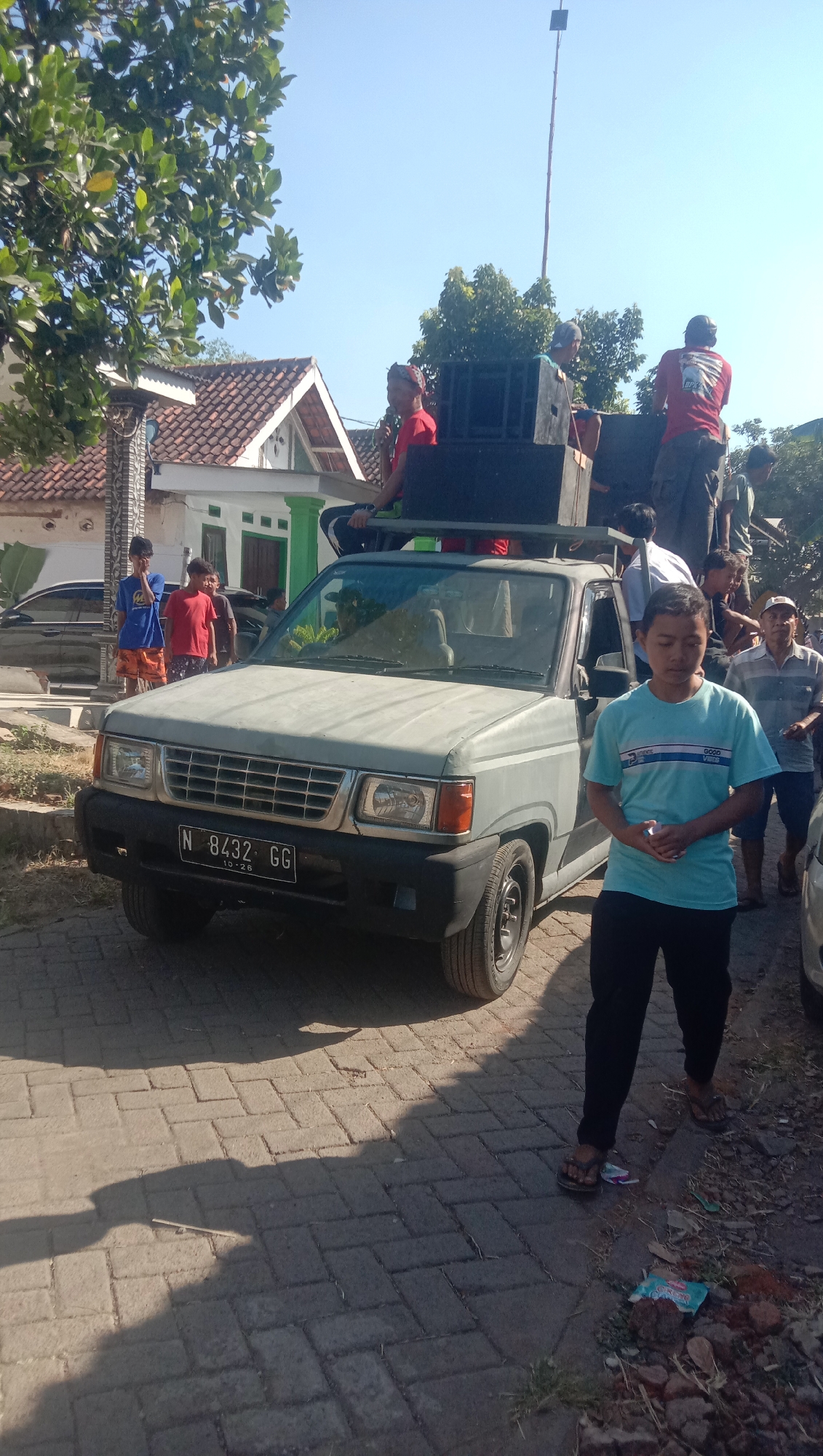 The image depicts a light blue pickup truck parked on a paved street, possibly in the city of Ngempit, Indonesia, under a clear blue sky. The truck is equipped with large speakers mounted on its roof and in its bed, suggesting it is being used for an outdoor event or mobile sound system. Several individuals are visible around and on the truck. In the foreground, a young person wearing a light blue t-shirt and dark pants stands facing away from the truck, looking down. Behind the truck, several people are standing and sitting, some appearing to be preparing or operating the sound equipment. One individual in a red shirt is seated on the edge of the truck bed. Further in the background, houses with tiled roofs and trees are visible, indicating a residential area. The street is paved with interlocking bricks, and shadows from the trees and people are cast on the ground.