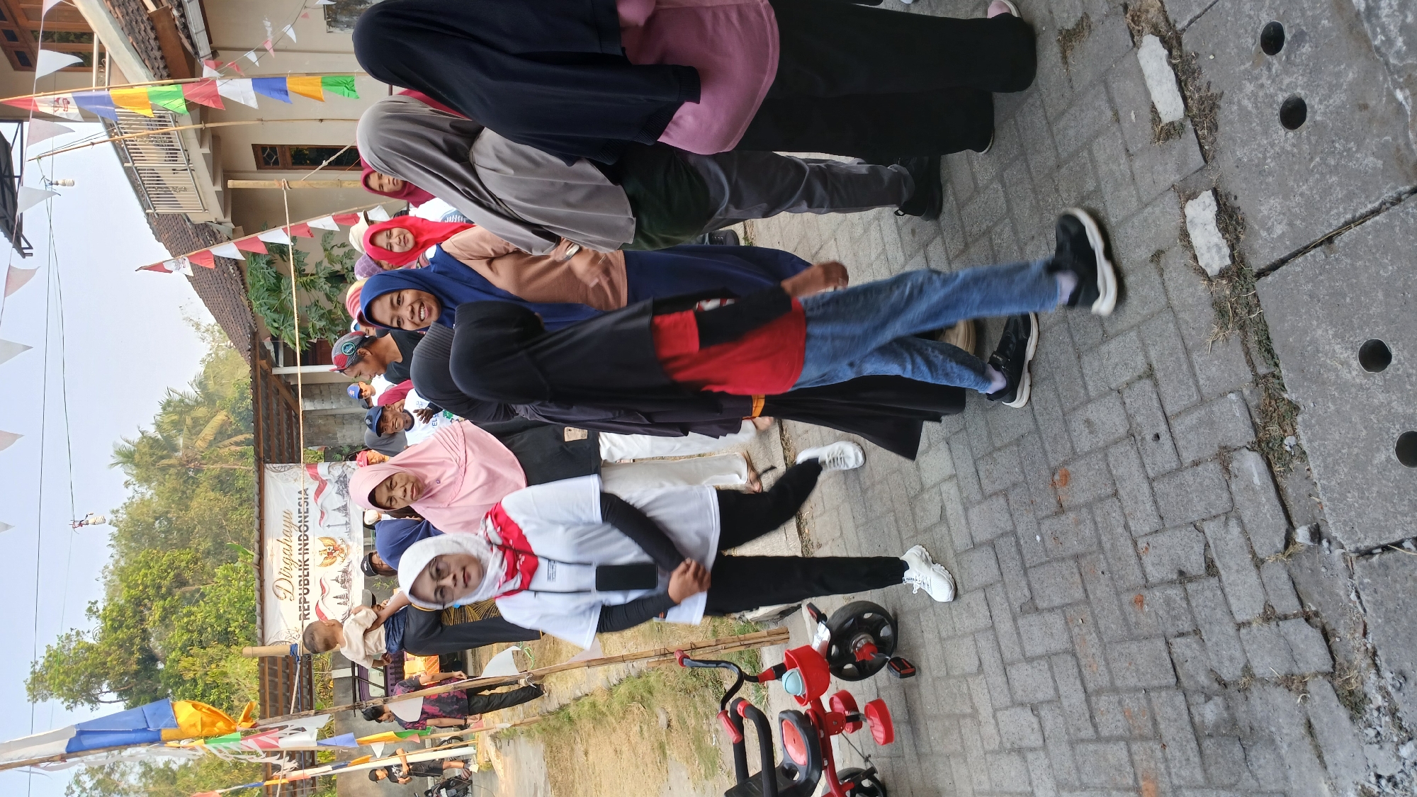 In the city of Mendut, Indonesia, a group of people are gathered on a brick-paved street, possibly for a community event. A banner above reads "Dirgahayu REPUBLIK INDONESIA," indicating a celebration of Indonesian Independence Day. Several individuals, mostly women, are wearing hijabs and modest clothing. A child is being held by an adult, while others are walking and standing. A red tricycle is parked to the left of the frame. The background shows buildings and trees, suggesting a residential area. The lighting indicates it is daytime, likely morning or late afternoon.