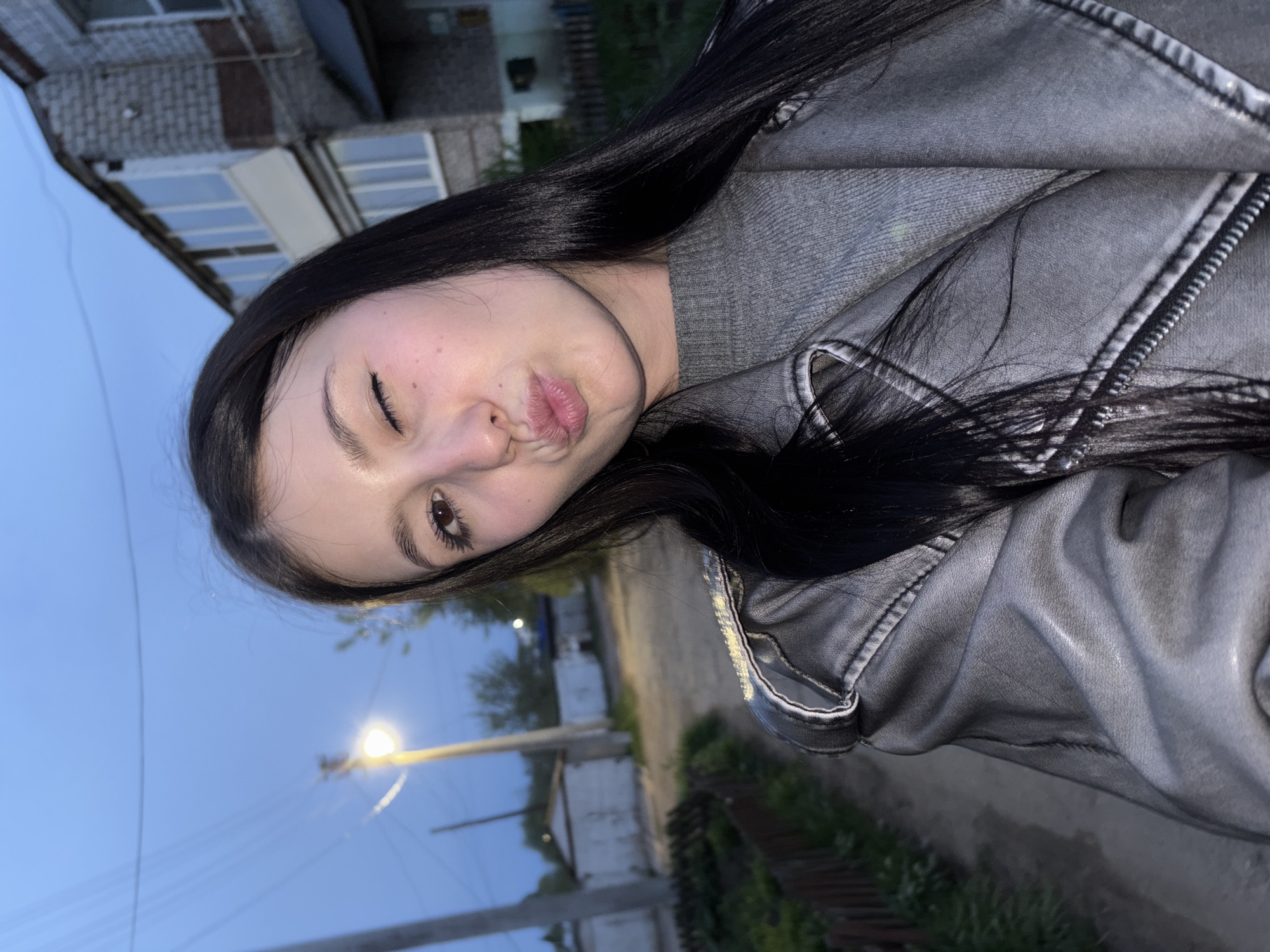 A young woman with long, black hair and fair skin stands outside in the city of Фоки, Russia. She is wearing a gray sweater and a gray denim jacket. She has her lips pursed in a kiss and is winking one eye. Behind her, a street is visible with a streetlight illuminating the scene. A building with windows and a brick exterior can also be seen in the background. A grassy patch and the edge of a sidewalk or path are visible at the bottom right. The sky is a dark blue, suggesting it is either dusk or dawn.