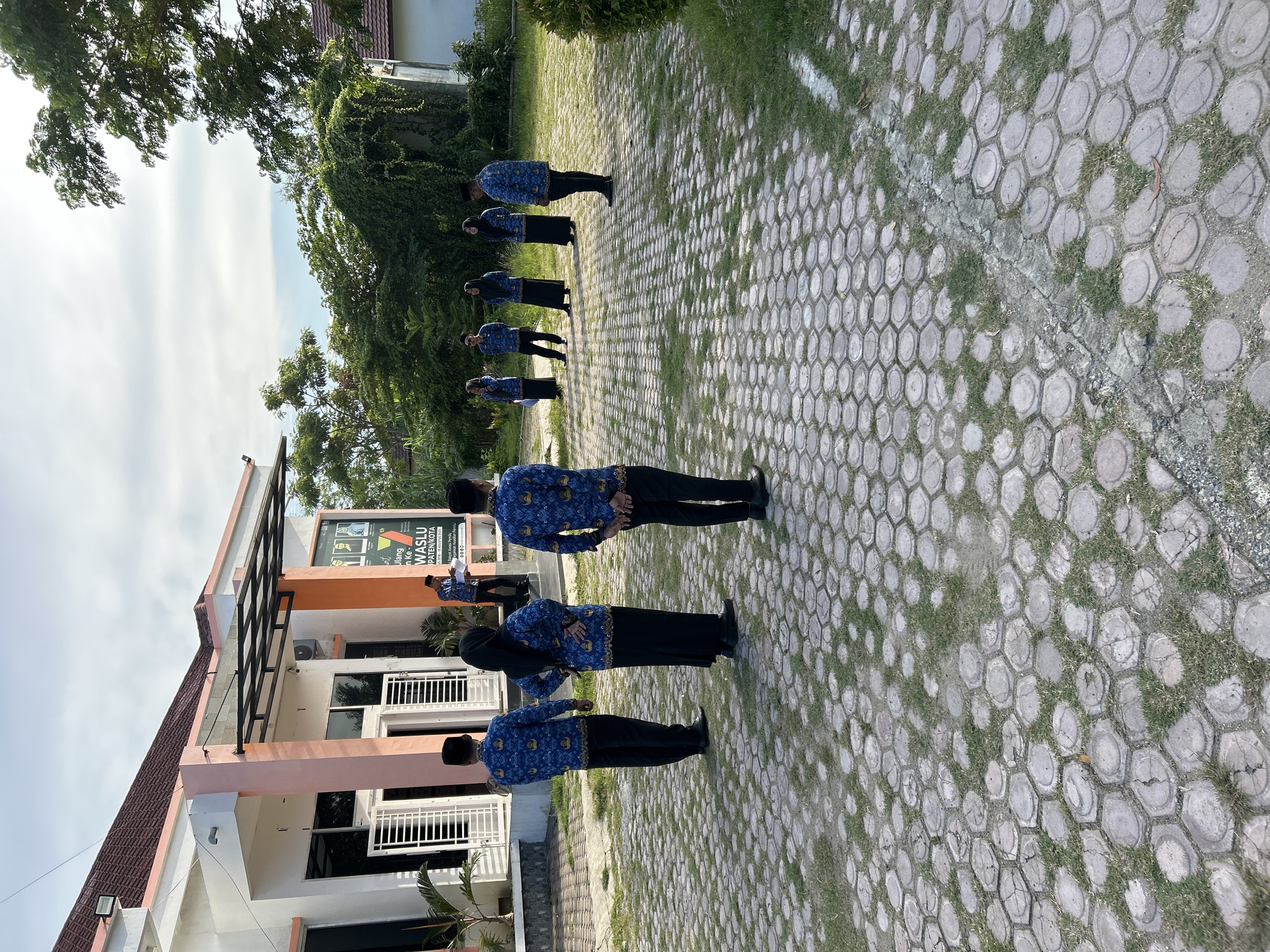 A group of people stand at attention in front of a two-story building. The building is white and peach-colored with black trim. A sign on the building reads "WASLU PATENKOYA" in a blue field. The people are wearing blue shirts with a repeating pattern and black pants or skirts. They are standing on a paved area made of hexagonal blocks with grass growing in the spaces between the blocks. A tree line is visible in the background. The location is Lhokseumawe, Indonesia.