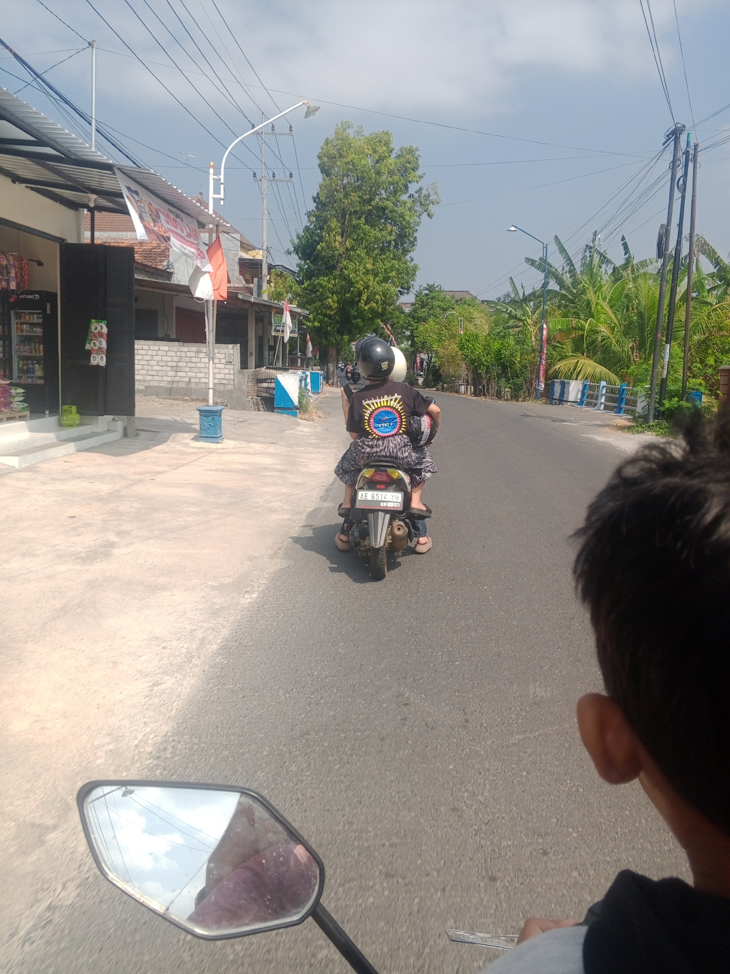 In the city of Ronowijayan, Indonesia, a daytime shot captures two individuals riding a scooter on a paved road. The person in the rear, wearing a black helmet and a black t-shirt with a circular graphic design, sits in front. They are accompanied by a second person, whose head and shoulder are visible in the foreground, looking forward. The scooter, a white model with a license plate reading "AE 6514 YH", is in motion, suggesting travel. The environment features buildings with corrugated metal roofs and a brick wall on the left, while the right side of the road is lined with lush green trees, including banana plants. Power lines are strung overhead, and a street lamp is visible. A blue and white railing runs along the right side of the road in the middle distance. The perspective suggests the image is taken from a second scooter.