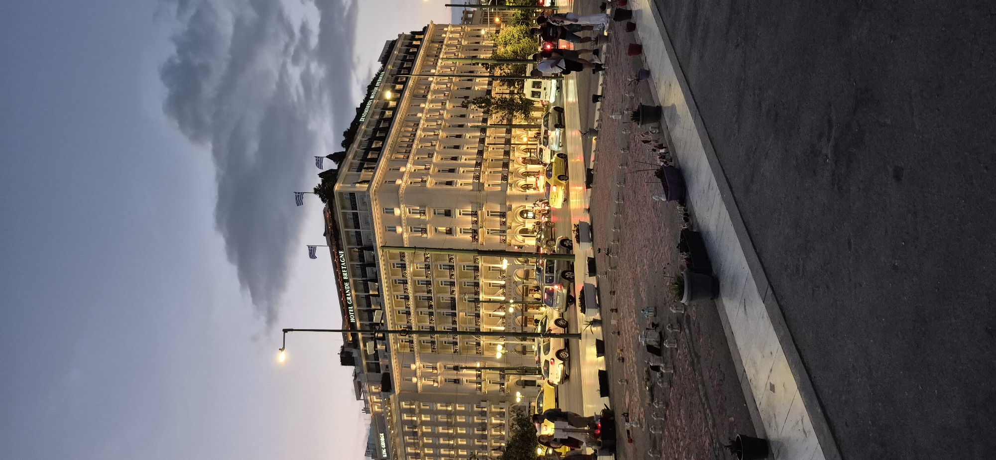 The image shows the exterior of the Hotel Grande Bretagne in Athens, Greece, at dusk. The hotel is a large, ornate building with multiple stories, balconies, and illuminated windows. Several Greek flags are visible on the rooftop. In front of the hotel, there is a street with several cars, including yellow taxis. On the sidewalk in the foreground, there are numerous small white candle-like objects arranged in a pattern, along with potted plants. A few people are standing on the sidewalk, some looking at the hotel or the memorial. The sky is a soft gradient of blue and grey, with some clouds visible. The overall scene suggests a calm evening in a busy urban setting, with a somber memorial present.