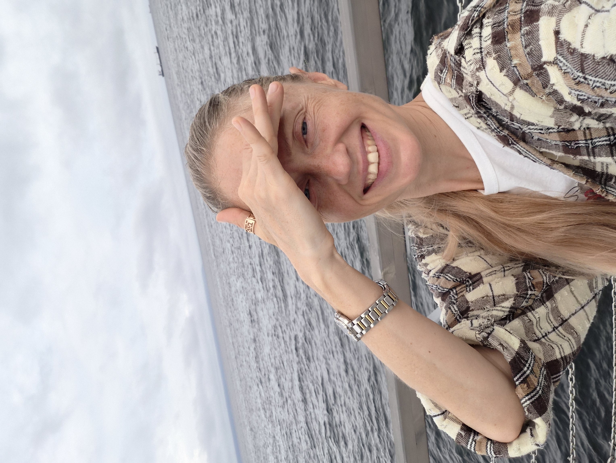 A Caucasian woman with fair skin, freckles, and blonde hair pulled into a ponytail is taking a selfie outdoors. She is wearing a white t-shirt and a plaid jacket. Her right hand is raised to her forehead, partially shielding her eyes from the sun. She is smiling, showing her teeth. A silver and gold watch is on her left wrist, and a gold ring is on her right index finger. The background shows a body of water with a large ship in the distance, suggesting a coastal or harbor location. The sky is overcast. The railing in the foreground indicates she is on a pier or boardwalk. Given the provided context of Vladivostok, Russia, this scene likely depicts a waterfront area within the city.
