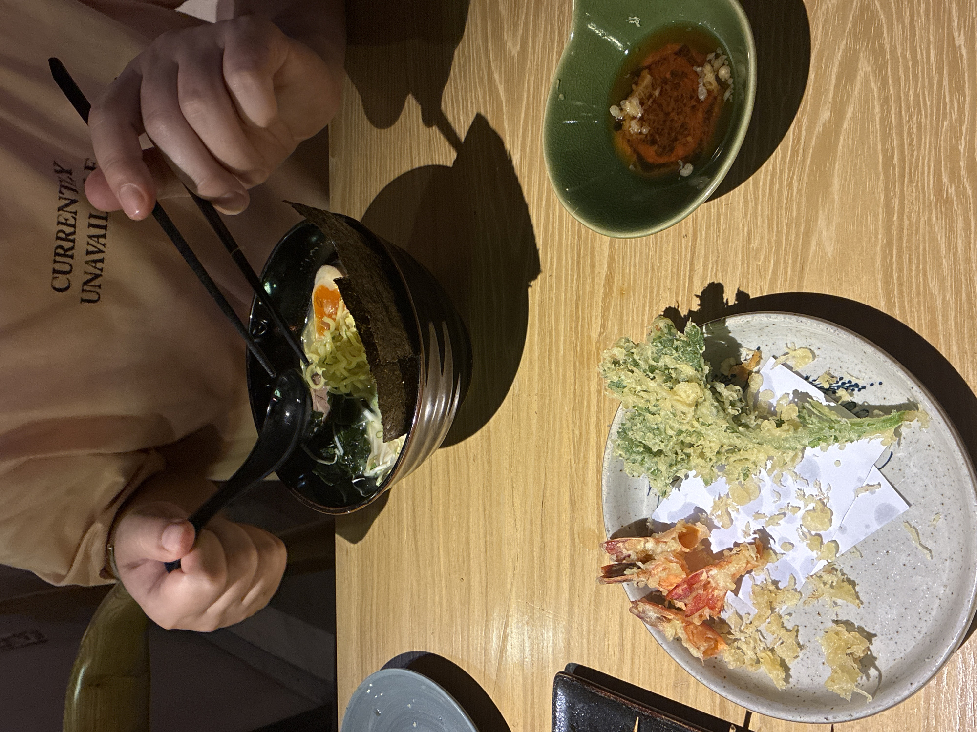 An aerial view shows a person seated at a wooden table, about to eat a meal. The person is using chopsticks to eat ramen, with a soft-boiled egg and seaweed in a black bowl. A small green bowl contains a dipping sauce, and a light gray plate holds shrimp and vegetable tempura with a sheet of white paper to absorb excess oil. The person is wearing a peach-colored shirt with lettering. The setting could be a restaurant in Đà Nẵng, Vietnam, though it's not possible to confirm this with certainty.