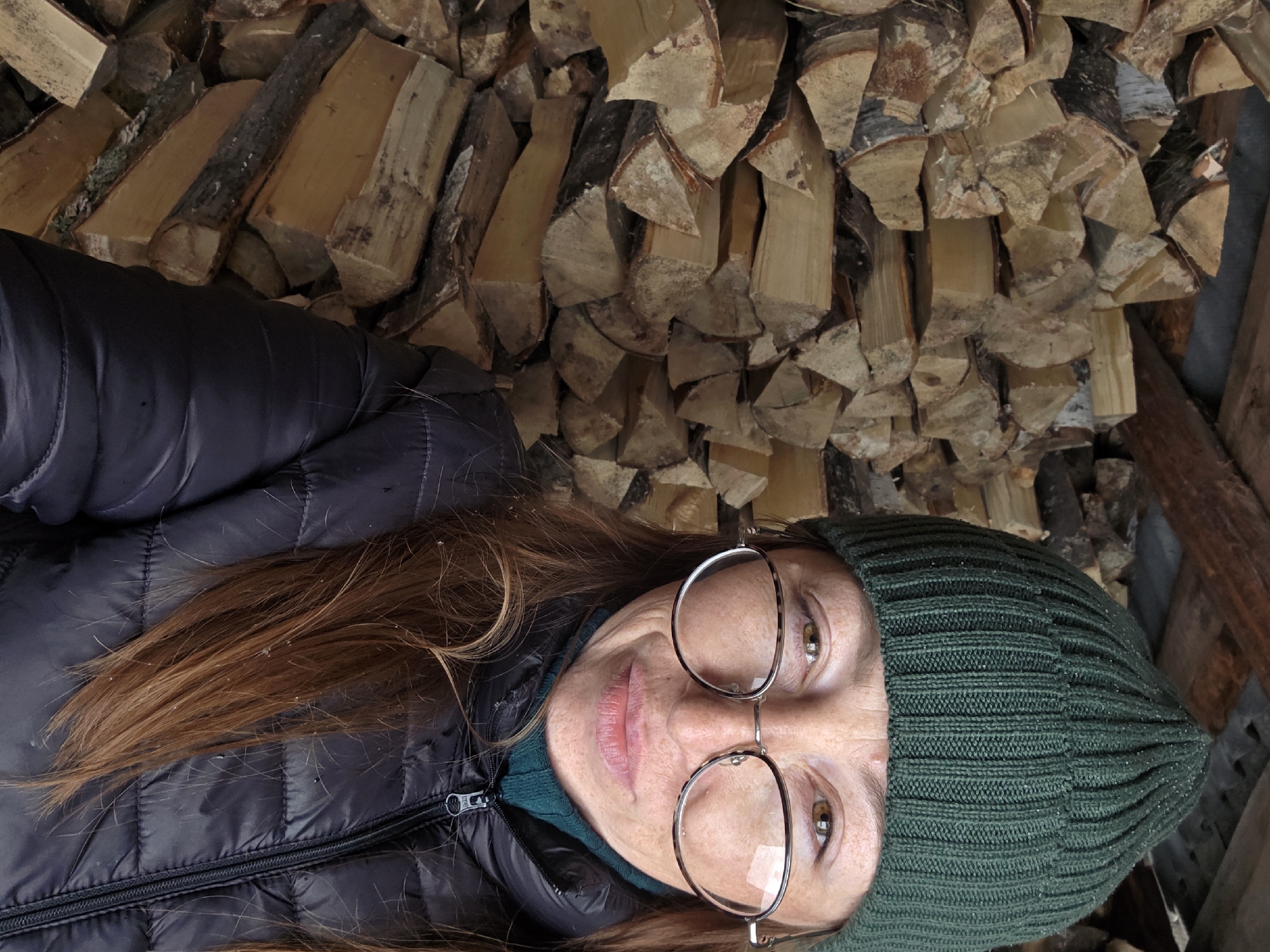 The image depicts a woman lying on the ground, with her head towards the lower part of the frame and her body extending upwards. She is wearing a black quilted jacket, a green beanie, and round eyeglasses. Her hair is auburn and falls over the jacket. Behind her is a stacked pile of cut firewood. The location is not specified and cannot be extracted from the image; the reference to Зипуново, Russia is extraneous.