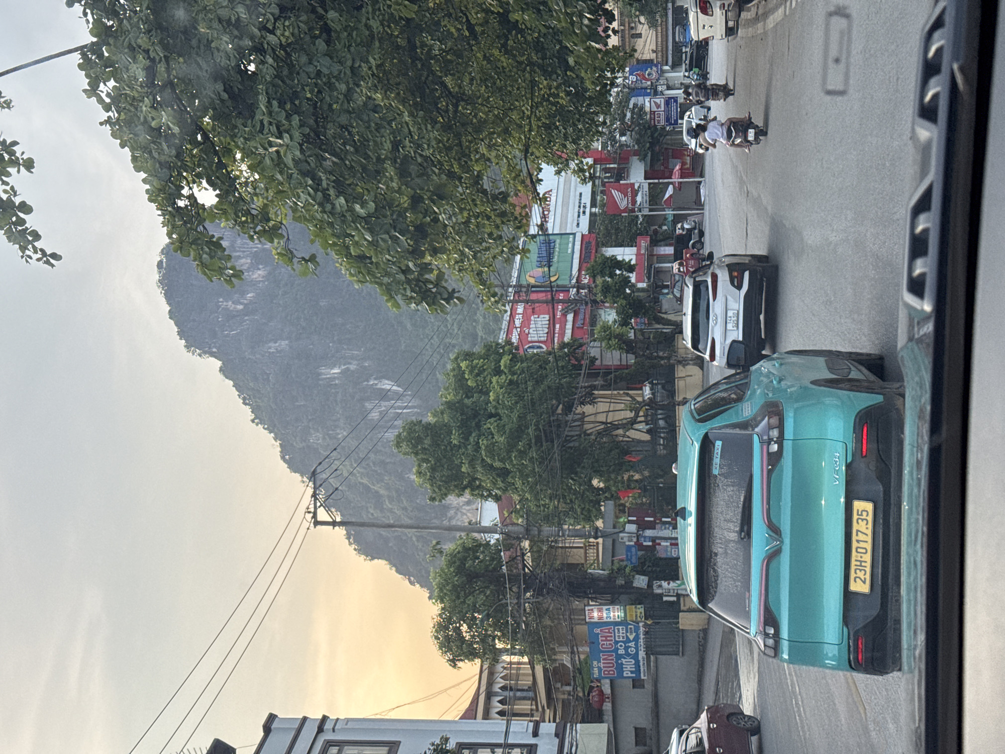 The image is a street scene in Hà Giang 1 Ward, Vietnam, showing a road with traffic, including cars, motorbikes, and pedestrians, lined with buildings, utility poles, and trees.
In the foreground, a turquoise VinFast VF e34 car with the license plate "23H-017.35" is visible. Traffic is moving on the right side of the road, including a white car with the license plate "14A 525.50", and several motorbikes. A building with a sign saying "Bún Chả Phở" is also visible. The area has numerous advertisements, including those for Honda.
In the background, a large, tree-covered mountain is seen against a hazy sky. The overall impression is of a busy urban environment with natural elements in the background.