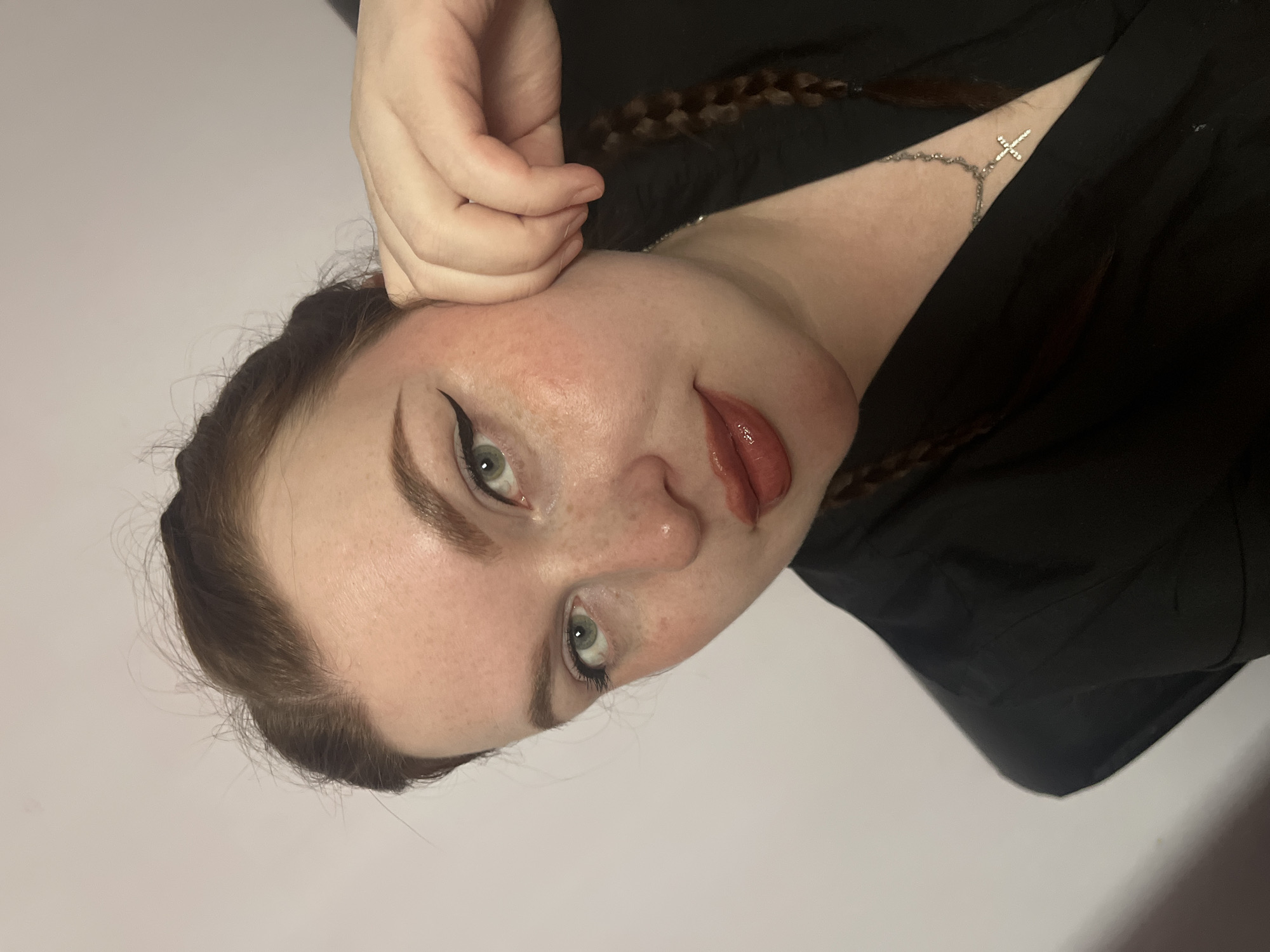 A fair-skinned person with braided reddish-brown hair and a silver cross necklace is pictured close-up against a plain white background. The person is oriented with their head tilted to the right. Their left hand is near their cheek. Their makeup includes black eyeliner. The image is likely taken indoors. There is no evidence to place this image in Kuyeda, Russia.