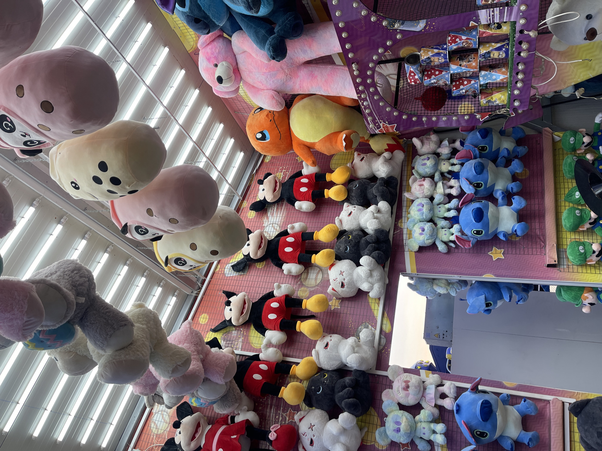 This is a low-angle, medium shot of a carnival game stall at the Varna Mall, Varna, Bulgaria. The stall is filled with various plush toys hanging from the ceiling and displayed on shelves. Prominent among the toys are several large Mickey Mouse plush figures, a large orange Charmander plush, a large pink teddy bear, and several Stitch plush toys in different sizes. There are also smaller plush toys of various characters hanging in rows. The background features bright fluorescent lighting. A prize shelf with stacked cups featuring cartoon characters is visible on the right side of the image. The overall scene depicts a colorful and inviting carnival atmosphere.