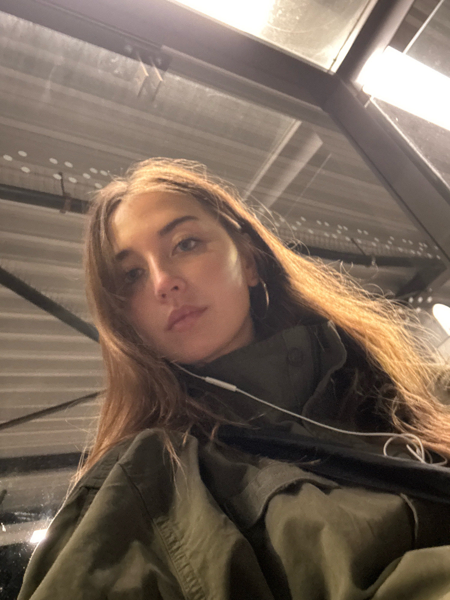 A low-angle, close-up selfie shows a young woman with long, brown hair and light brown eyes. She is wearing a dark green jacket, and a white wire for earbuds is visible. Her facial expression is neutral, and her gaze is directed slightly upwards and to the left. The background consists of a corrugated metal ceiling and fluorescent lighting, suggesting an indoor, possibly industrial or utilitarian setting such as a parking garage or a covered walkway. No specific location in Amsterdam is identifiable from the visual cues.