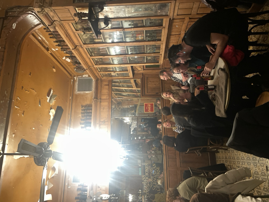 A crowded bar with a mix of people seated and standing around tables enjoying drinks. The interior features wooden paneling, mirrored walls, and glass cabinets filled with bottles. A Coca-Cola sign is visible. The ceiling has ornate molding, with an old-fashioned ceiling fan. The walls are peeling. The overall atmosphere appears lively and historical.