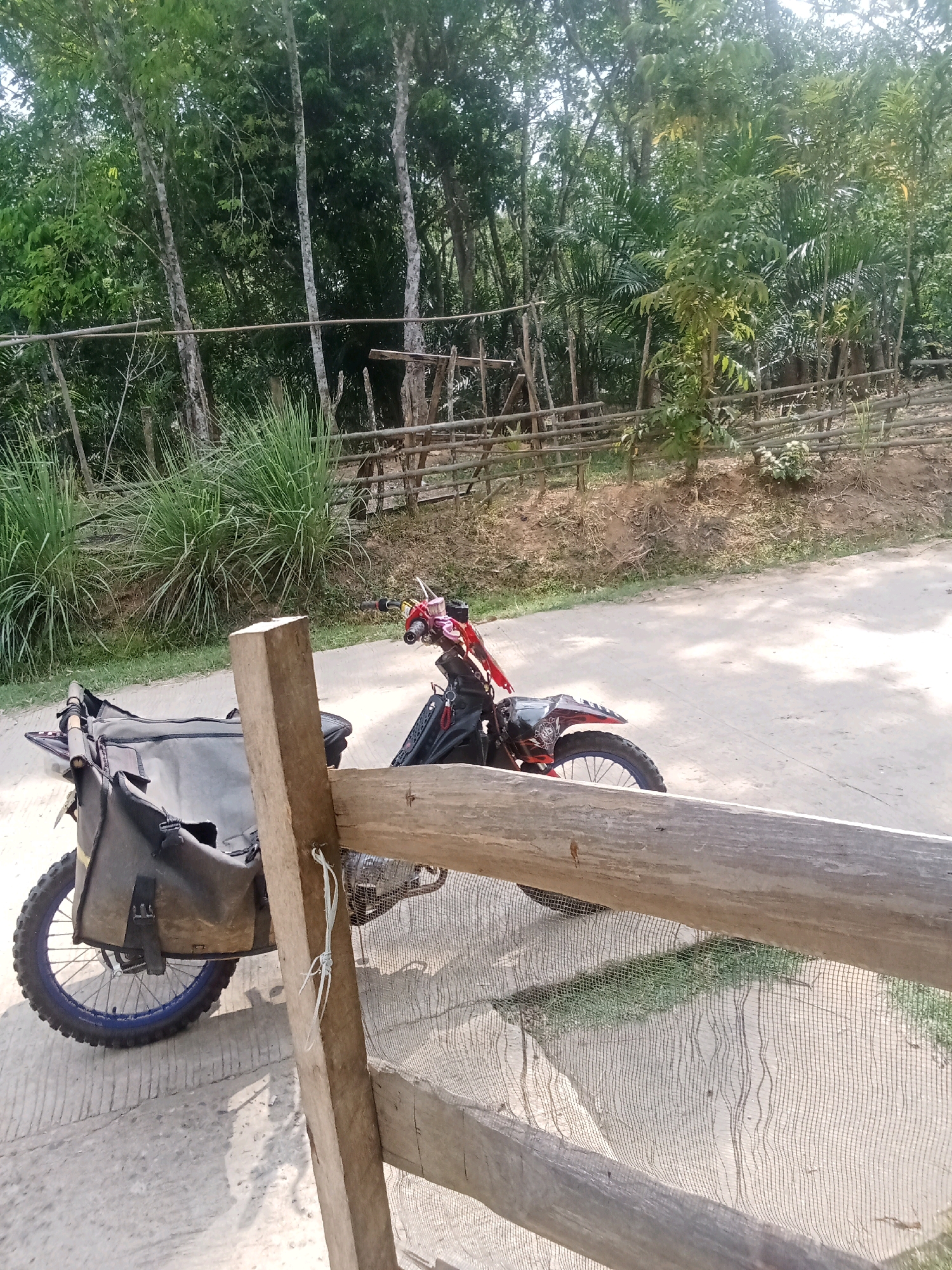 A dirt bike is parked on a dirt road next to a wooden fence. The bike is red and black and has a large black bag attached to the back. The fence is made of rough-hewn wooden planks and is partially covered in netting. Behind the fence, there are trees and vegetation. The scene appears to be in a rural area of Jirak, Indonesia. The lighting suggests it is daytime.