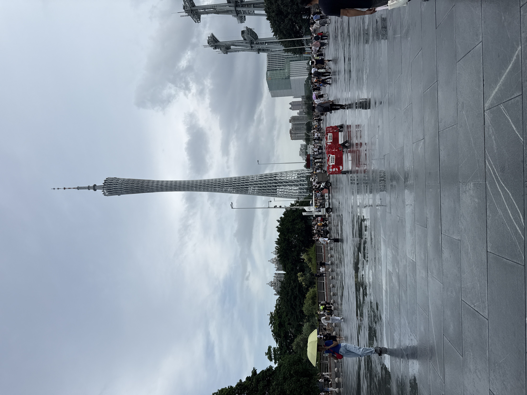 In Tianhe District, China, on a wet day with a cloudy sky, a wide, dark-tiled plaza reflects the surrounding environment. In the background, the prominent Canton Tower, a lattice-like structure, stands tall against the sky. To its right, two large, industrial-looking towers with intricate metalwork are visible. A crowd of people is scattered across the plaza, some holding umbrellas, indicating the wet weather. In the foreground, a woman with a yellow umbrella walks away from the camera, wearing light blue jeans and a dark top, carrying a red bag. The plaza is wet, with visible reflections of the sky and people. Several red barriers are placed on the ground, with Chinese text written on them. In the mid-ground, there are trees and buildings. A white vehicle, possibly a tour bus or tram, is also present. The overall atmosphere is one of a public space with moderate pedestrian activity on a damp day.
