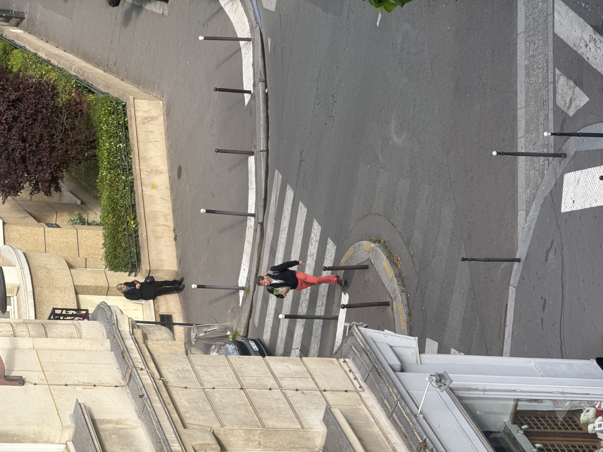A high-angle shot shows a street scene in Paris, France. A crosswalk bisects a grey asphalt street lined with rows of metal bollards and narrow concrete curbing with yellow and white markings. Two people are visible: one is crossing the crosswalk, wearing a dark jacket and bright red pants, carrying something green in their hands. The second person is lying on their side on the sidewalk next to a building and a planting bed filled with low, green shrubbery. The buildings are made of tan stone and white concrete, with visible architectural details. A parked silver car is also seen on the street.