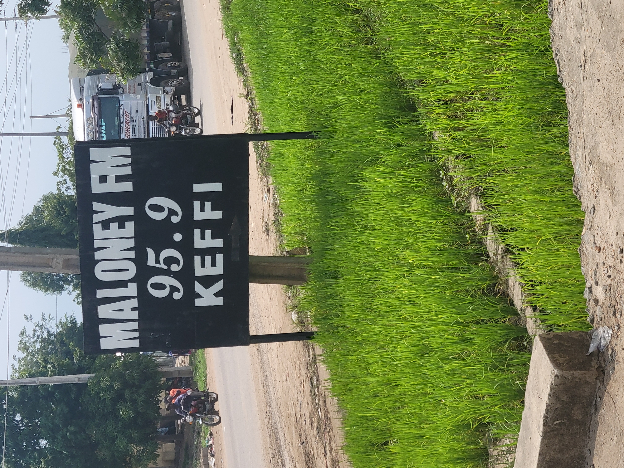 The image shows a black sign with white text that reads "MALONEY FM 95.9 KEFFI". The sign is mounted on a pole next to a road in Keffi, Nigeria. In the background, a large silver truck with a trailer is driving down the road. A person on a motorcycle is also visible in the distance, as well as some trees and buildings. The foreground of the image is filled with lush green grass. The scene appears to be a typical roadside view in a semi-urban or rural area of Keffi, Nigeria.