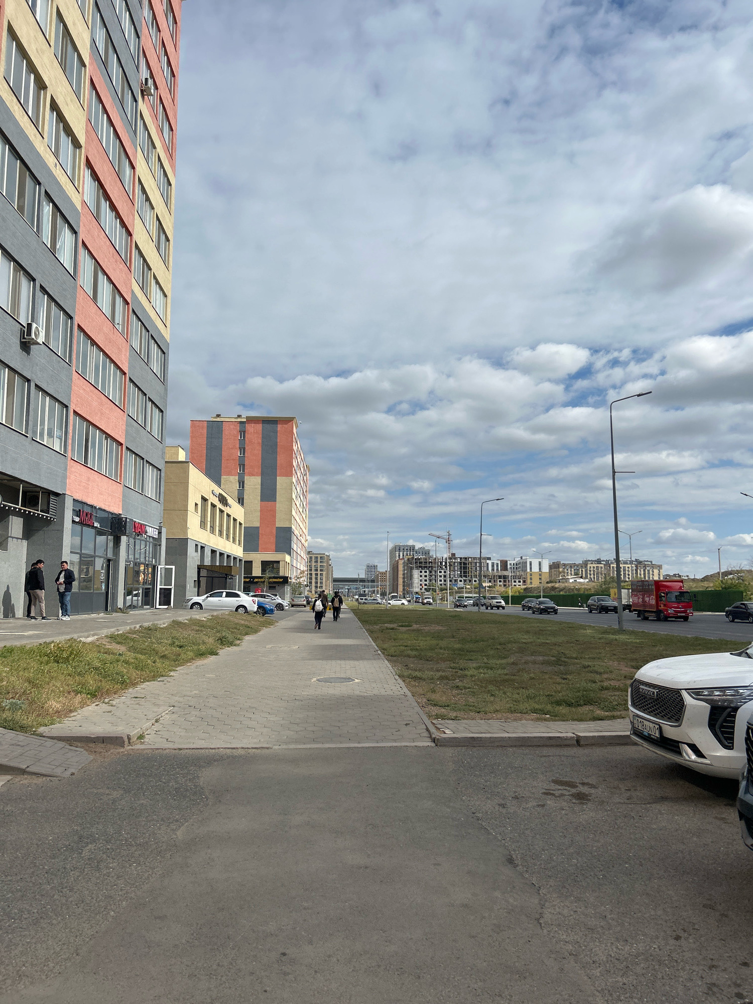 This is a street-level, vertical, full-shot photograph taken in Astana, Kazakhstan. On the left, a tall, modern apartment building with sections of grey, pink, and yellow facade dominates the foreground. The building features numerous windows and outdoor air conditioning units. A sidewalk runs along the base of the building, with a few individuals standing and walking on it. To the right of the sidewalk is a strip of dry grass and then a street with several cars driving and parked. In the background, more apartment buildings and construction cranes are visible, indicating ongoing urban development. The sky is filled with scattered white clouds against a pale blue background. A red truck with "Coca-Cola" branding is visible on the street in the midground.