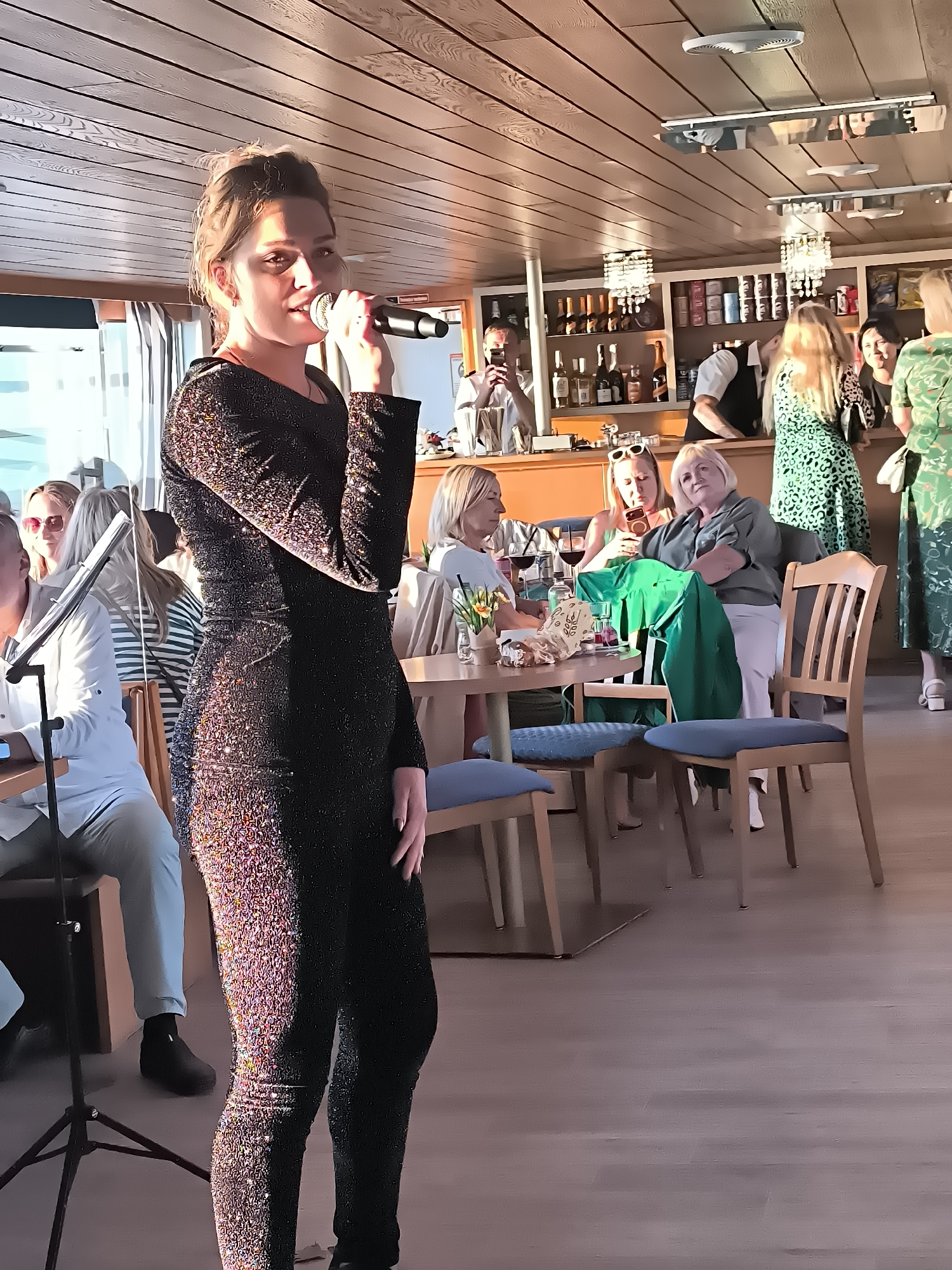A female singer is performing in a dimly lit establishment with a bar in the background and several patrons seated at tables. She is wearing a sparkling black outfit and holding a microphone. The environment appears to be a restaurant or bar, possibly on a boat given the wooden ceiling and the overall ambiance. The patrons are casually dressed and some are engaged in conversation. The lighting is warm and intimate. The location is the city of Neringa, Lithuania.