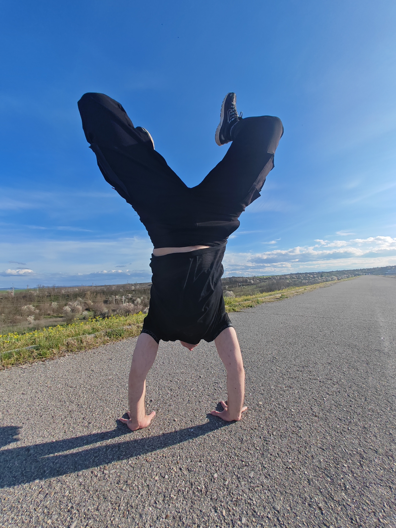 A person is performing a handstand on an asphalt road. The person is wearing black pants, a black t-shirt, and sneakers. The background includes a blue sky with some clouds, a grassy area with yellow flowers, trees, and buildings in the distance. The scene appears to be photographed outdoors on a sunny day. This picture was taken in Shumen, Bulgaria.