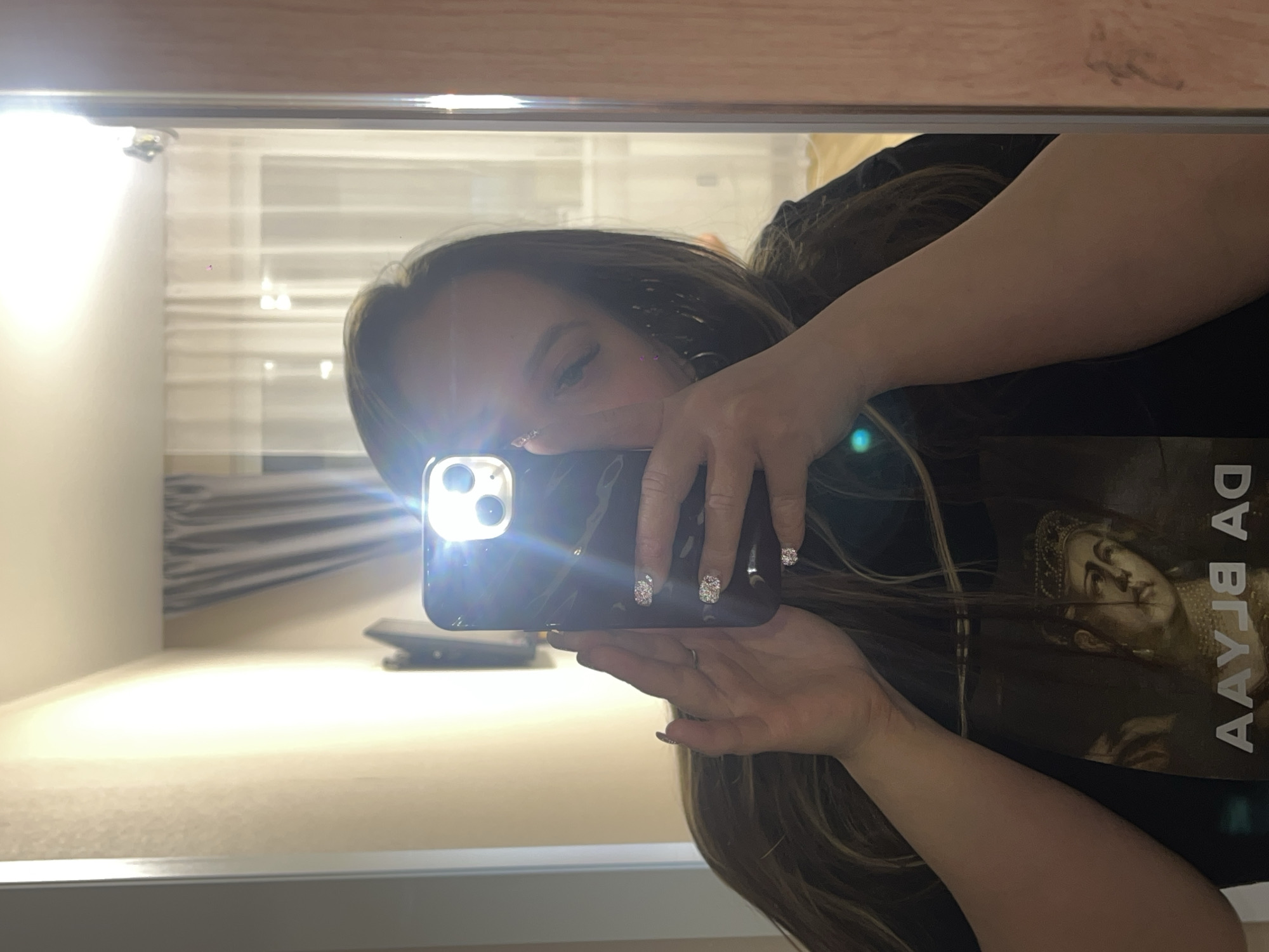 An indoor mirror selfie shows a person, likely female, holding a black smartphone with its flash activated, causing a bright glare that partially obscures her face. The image is rotated 90 degrees clockwise.
**Subjects:** The primary subject is the person reflected in the mirror, with long dark hair, wearing a dark t-shirt featuring a classical or historical portrait and text. Her hand holds the phone, showcasing nails decorated with glitter or rhinestones, and a ring on one finger.
**Objects:** The central object is the smartphone, likely an iPhone due to the camera lens layout, with a prominent flash. Other visible objects in the reflected room include light-colored walls, a window with light-colored blinds or sheer curtains, and a darker curtain. A surface or furniture piece is visible below the mirror's reflection.
**Scene/Setting:** The scene is an indoor room, possibly a bedroom or hotel room. The overall impression is well-lit due to the flash and reflected ambient light.
**Activity:** The person is engaged in the act of taking a mirror selfie.
**Notable details:** The phone's flash is very bright, creating lens flares and strong illumination. The person's face is largely hidden behind the phone and the intense flash.
**Visible Text:** On the person's dark t-shirt, text is visible in reflection, appearing reversed. It includes the letters "AЯL AD" and other distorted letters, alongside a depicted classical-style portrait of a person wearing a crown or headpiece. The presence of the Cyrillic letter "Я" suggests the text may be in Russian, which aligns with the provided location context of Krasnodar, Russia.