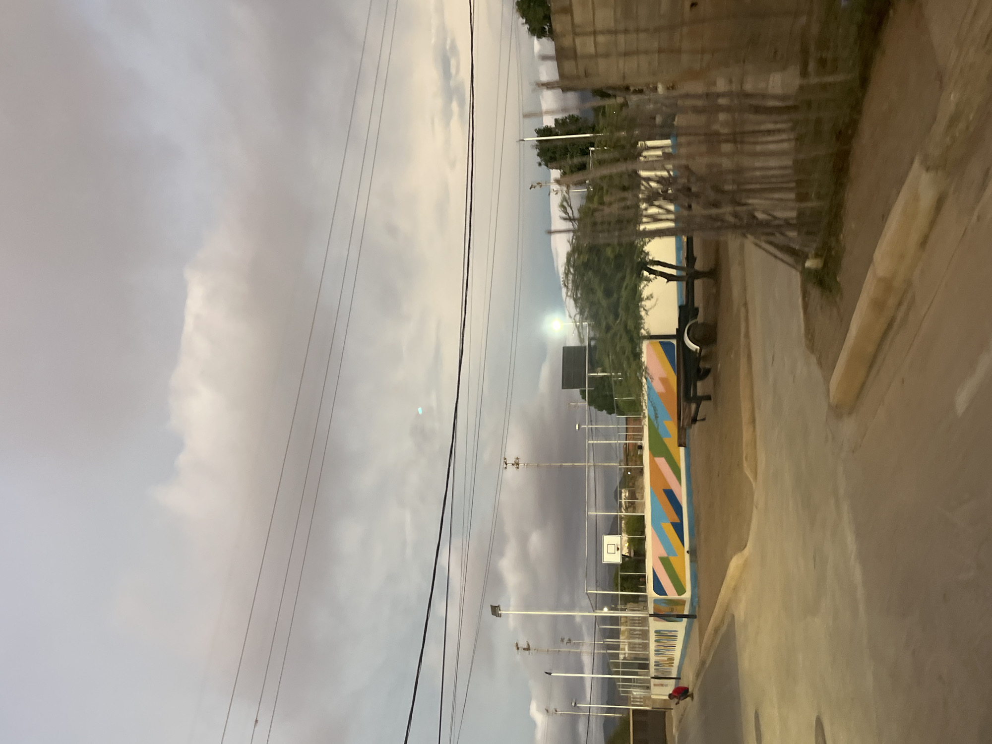 A cloudy evening in Juan Griego, Venezuela, reveals a colorful basketball court bordering a street. The court's outer wall displays geometric designs, juxtaposed against the utilitarian poles and wire mesh fence. An adjacent sidewalk leads past a modest wooden fence. Power lines stretch across the sky. A single individual is visible on the street.