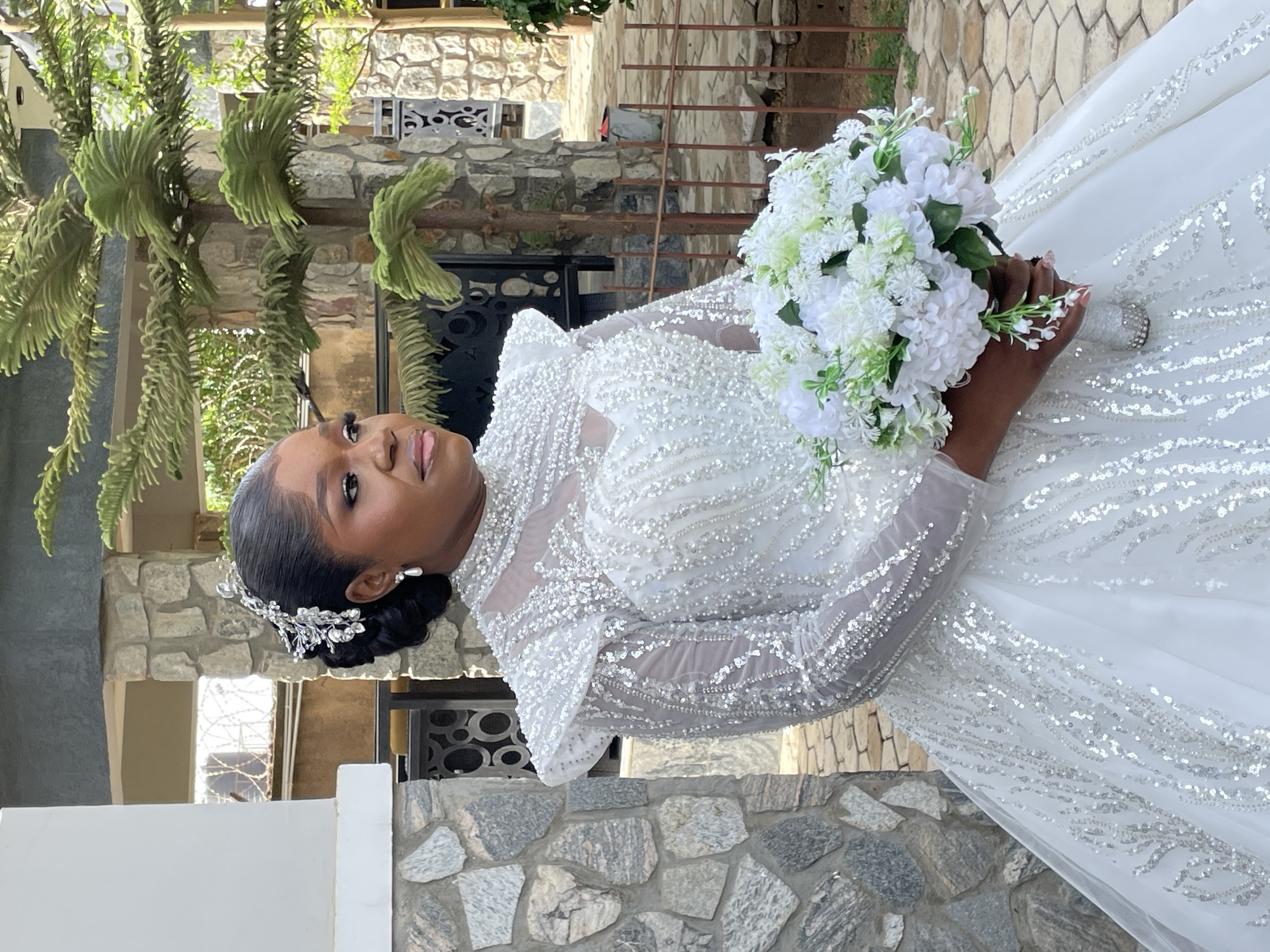 A woman is holding a bouquet of white flowers and wearing a white, long-sleeved wedding gown with silver embellishments. She is wearing a silver headpiece and earrings, and her hair is styled up. The background includes a building with stone walls, a metal gate, and plants. The image was taken outside, and the orientation is portrait. The location is Bukuru, Nigeria.