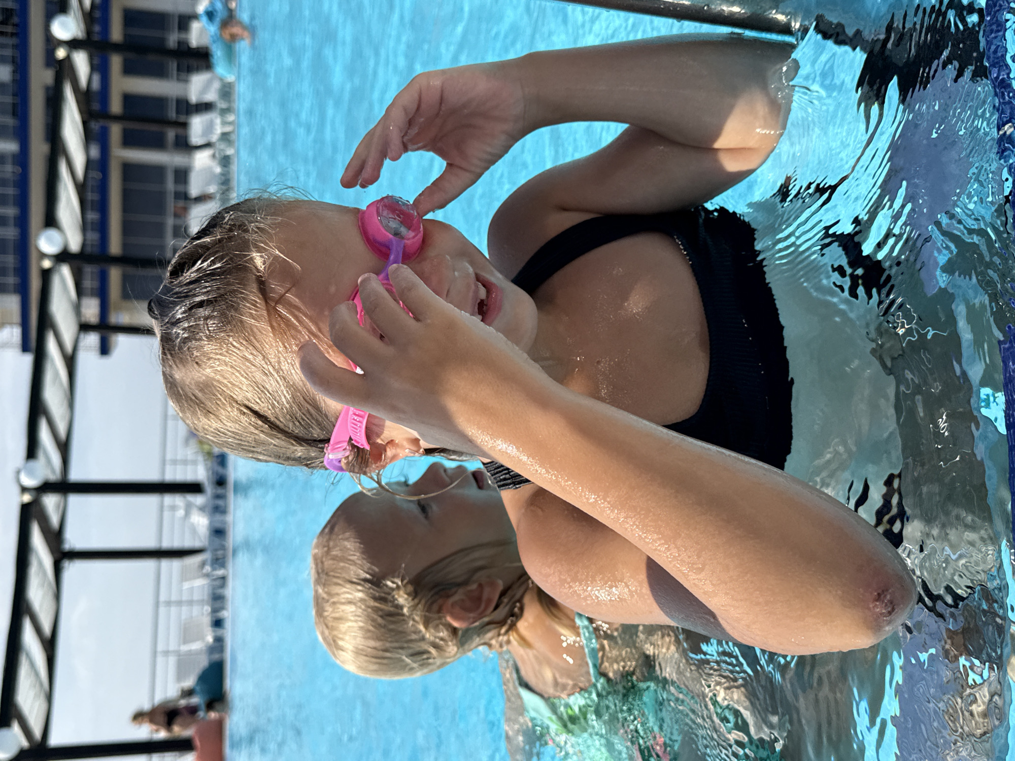 In a swimming pool in Sevastopol, Ukraine, a young girl wearing pink goggles and a dark swimsuit is holding her goggles up with her hands. Another child, with blonde hair, is partially visible behind her in the water. The pool is surrounded by lounge chairs and a building with a patterned roof. The water is a bright blue, and sunlight reflects off its surface.